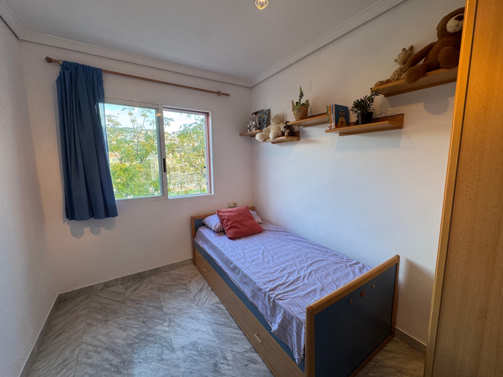 Einzelzimmer mit Marmorboden, Wandregalen aus Holz und Schiebefenster mit Blick ins Grüne. Das Bettgestell ist mit praktischen Schubladen ausgestattet.
