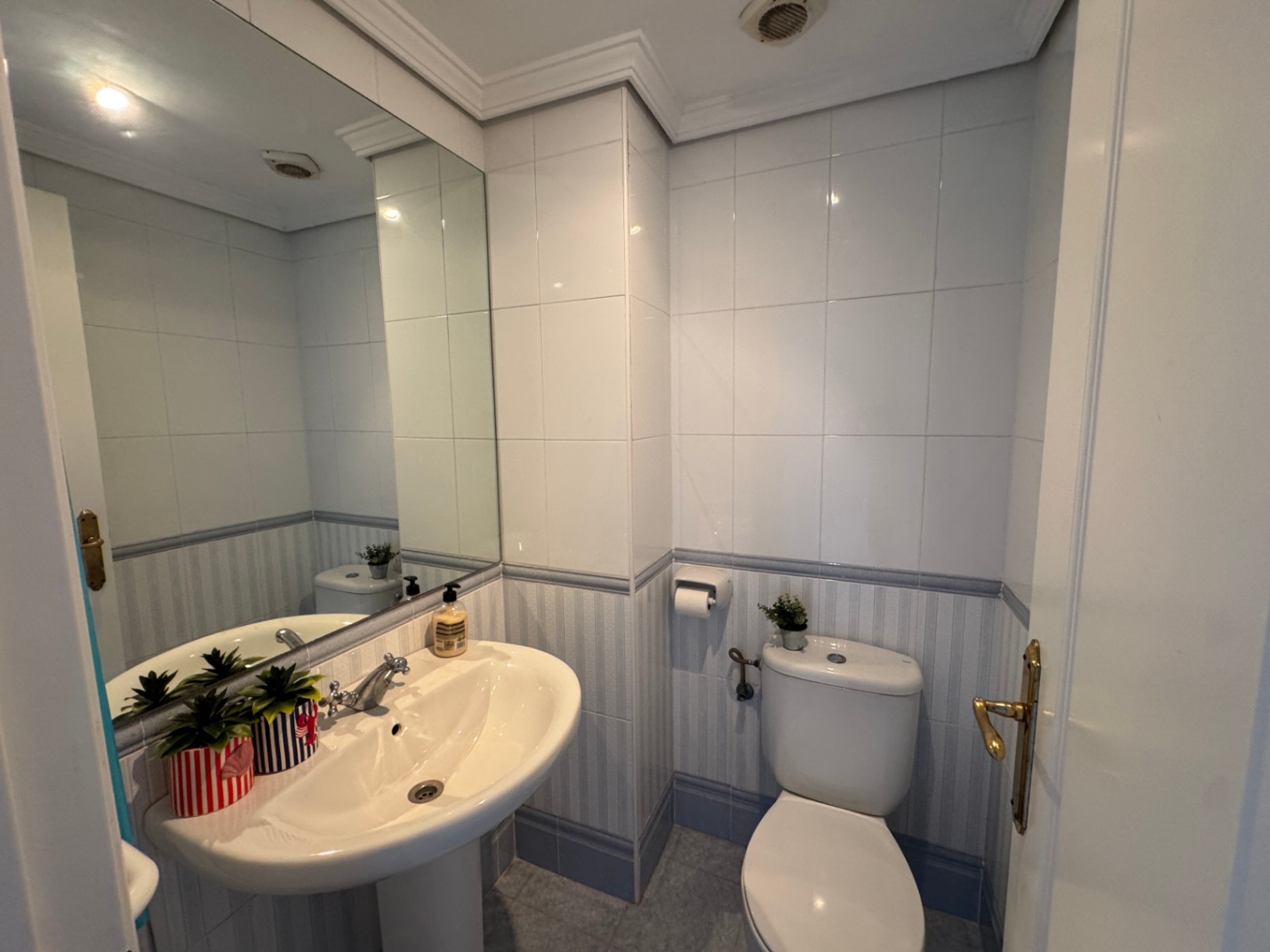 Funktionales Badezimmer mit Säulenwaschbecken, großem Spiegel und gefliesten Wänden mit einer blau gestreiften Bordüre.