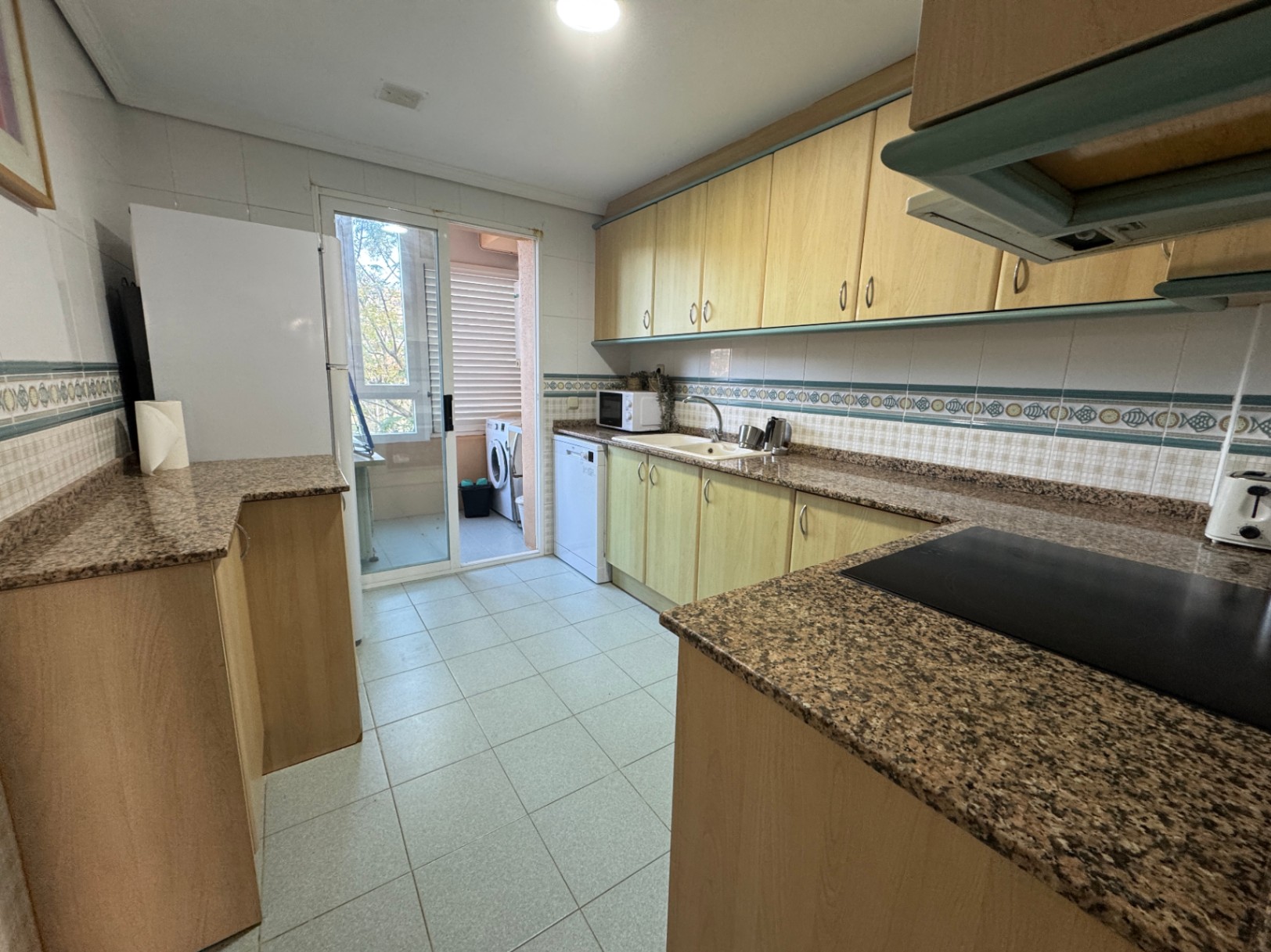 Küche mit hellen Holzschränken, Granitarbeitsplatten und Zugang zu einem Hauswirtschaftsbalkon mit Waschmaschine durch eine Glasschiebetür.