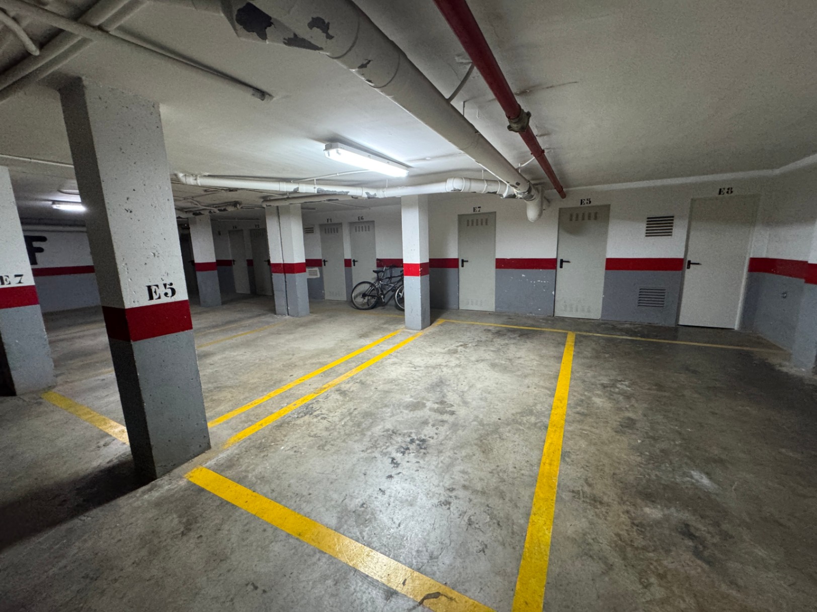 Tiefgarage mit Betonboden, gelben Markierungen und nummerierten Kellerräumen. Sichtbare Deckenrohre und funktionale Beleuchtung prägen den Raum.