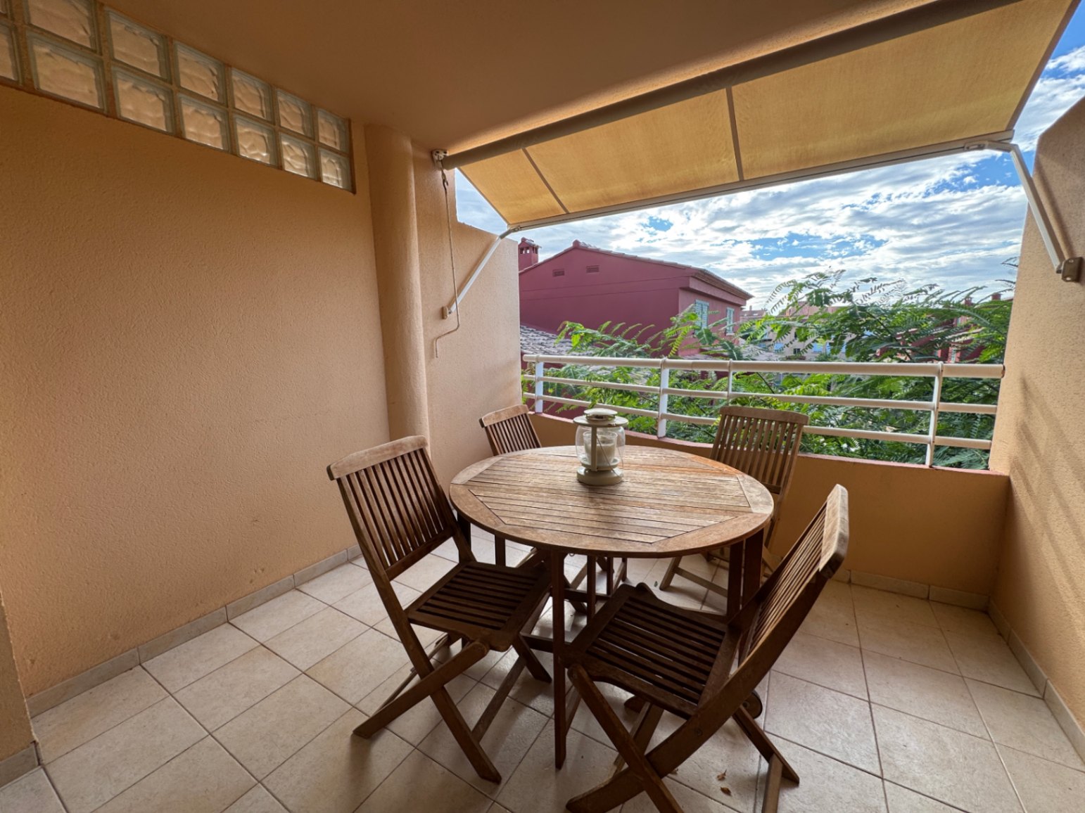 Überdachte Terrasse mit runder Holz-Sitzgruppe, Fliesenboden und ausziehbarer Markise mit Blick auf die umliegende Vegetation und Wohngebäude.