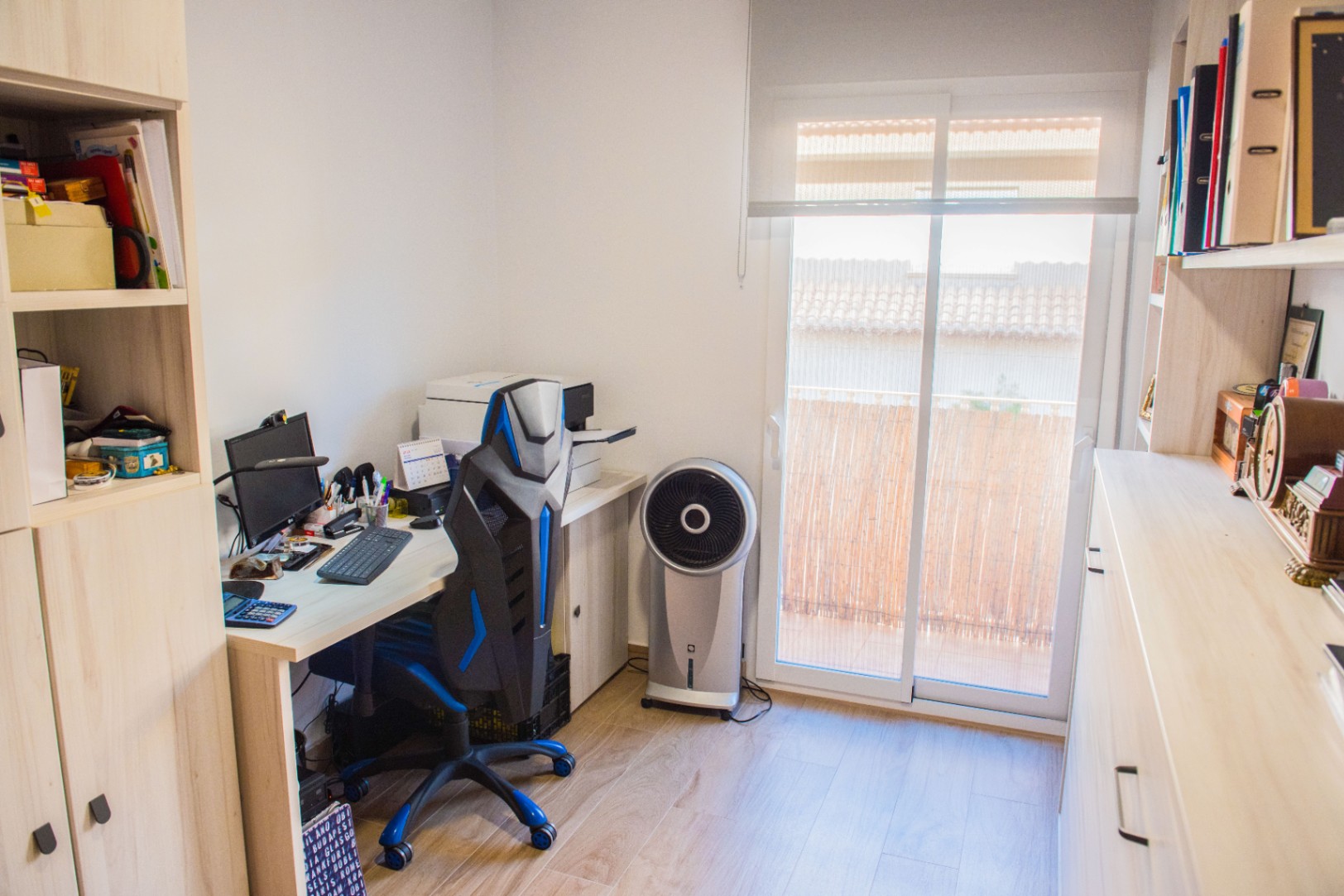 Arbeitszimmer mit hellem Holzschreibtisch, ergonomischem Bürostuhl und Einbauschränken. Eine Glasschiebetür bietet Tageslicht und Zugang zu einem Balkon mit Sichtschutz.