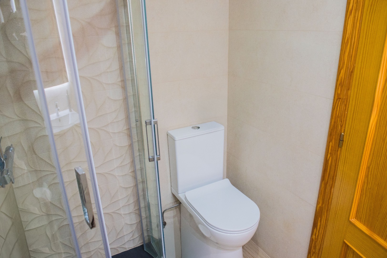 Badezimmerecke mit weißem WC und Glasduschkabine, akzentuiert durch strukturierte Wandfliesen mit Blattmuster und verchromten Griffen.
