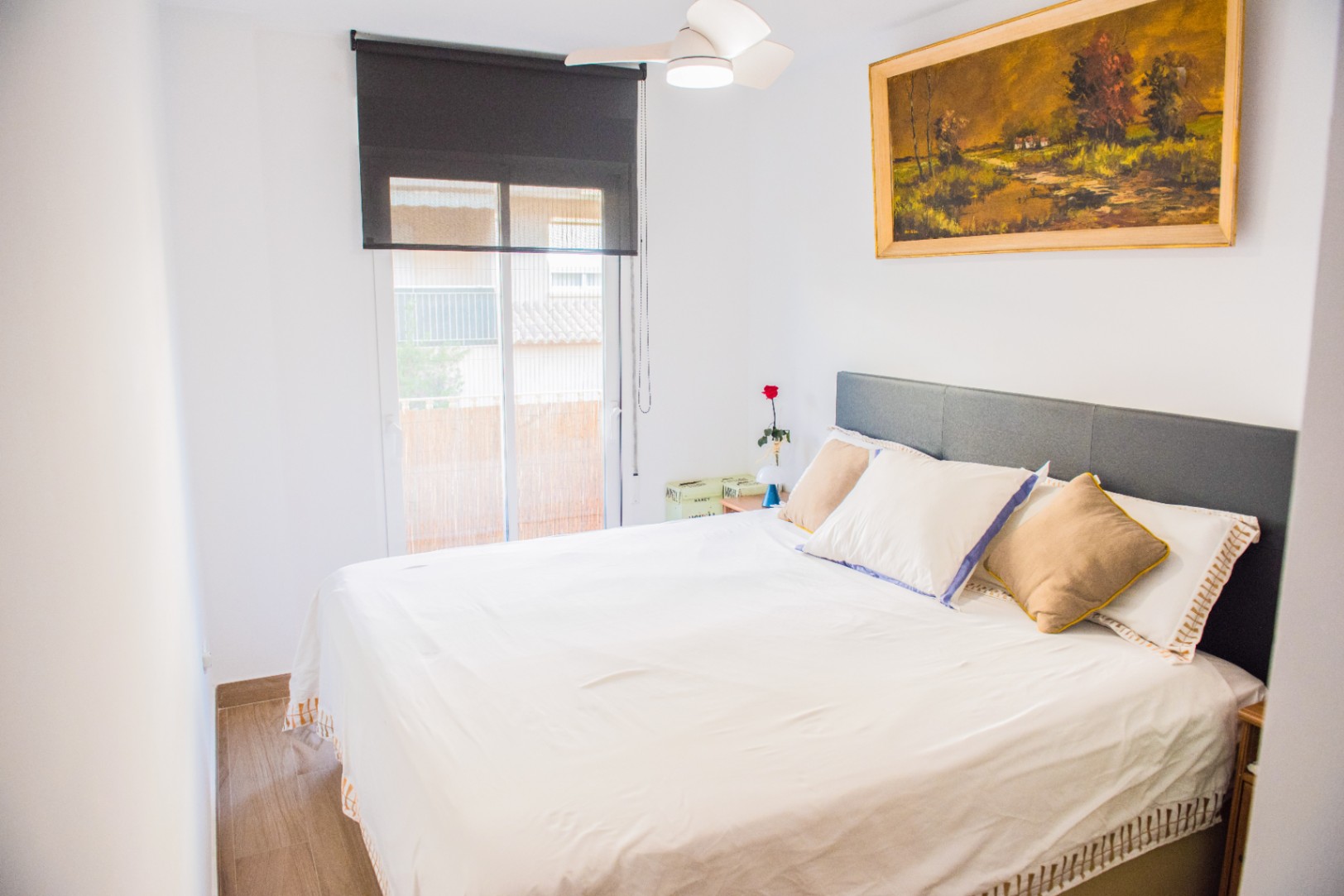 Helles Schlafzimmer mit grauem Kopfteil, Deckenventilator und schwarzem Rollo. Ein Ölgemälde mit Landschaftsmotiv ziert die Wand über dem Bett.