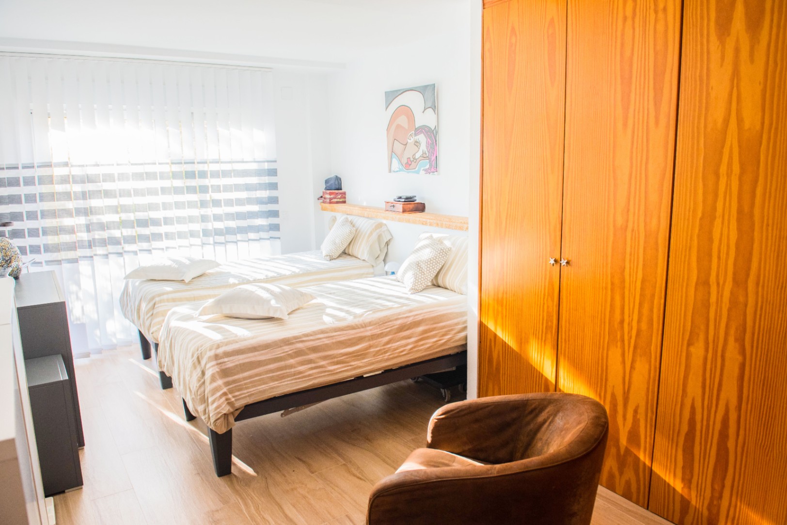 Helles Schlafzimmer mit zwei Einzelbetten, einem großen Einbauschrank aus Holz und einem Ledersessel auf hellem Laminatboden.