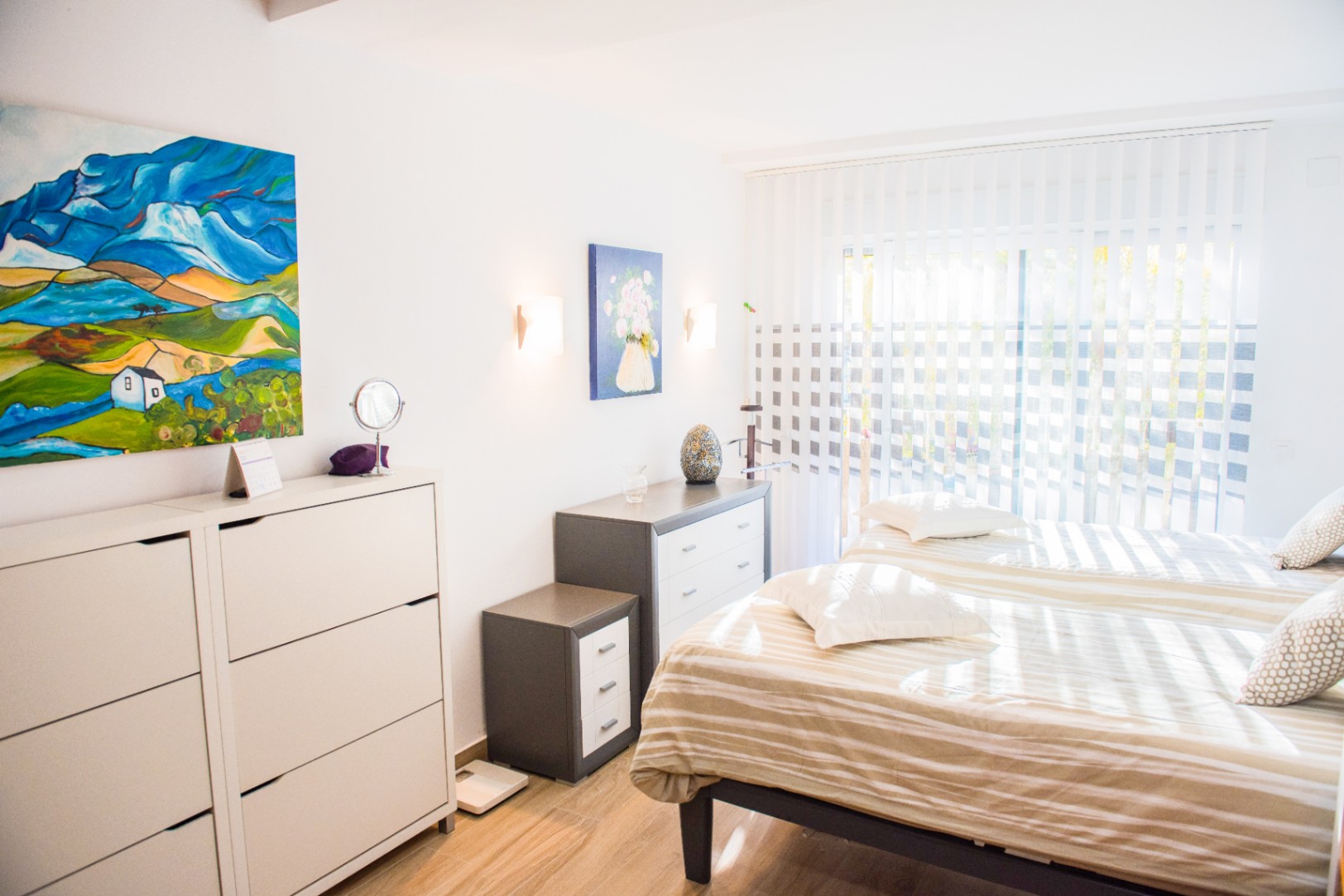 Helles Schlafzimmer mit zwei Einzelbetten, minimalistischen Kommoden und farbenfroher Kunst. Natürliches Licht fällt durch vertikale Lamellen auf den Boden in Holzoptik.