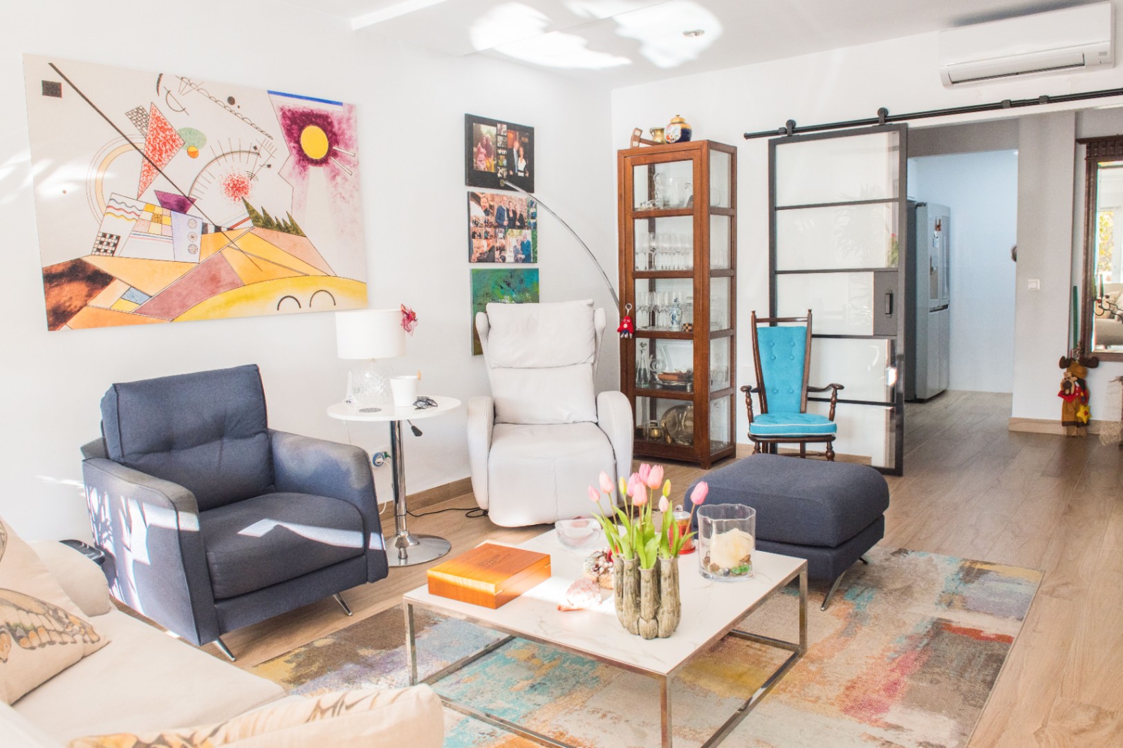 Helles Wohnzimmer mit blauem Sessel, weißem Relaxsessel und abstrakter Kunst. Eine Glasschiebetür mit schwarzem Rahmen trennt den Raum vom Flur.