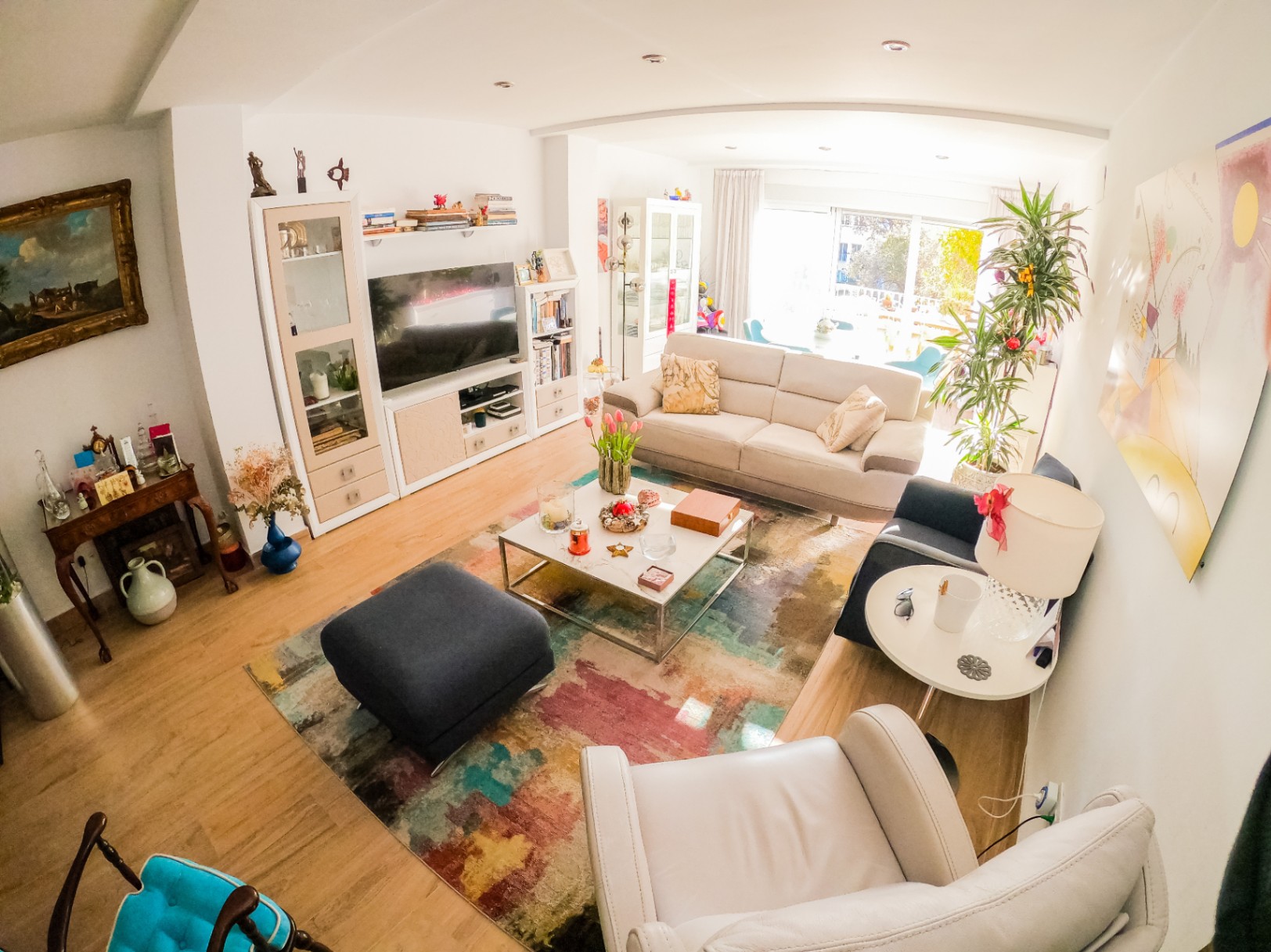 Helles Wohnzimmer mit hellem Holzboden, einem bunten abstrakten Teppich und modernen Möbeln. Große Glasschiebetüren bieten viel Tageslicht und Zugang zum Balkon.