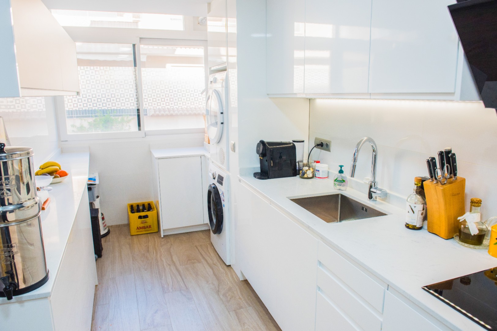 Moderne Pantryküche mit grifflosen weißen Schränken, integriertem Waschbereich und weißer Arbeitsplatte. Mit LED-Unterbaubeleuchtung und hellem Boden in Holzoptik.