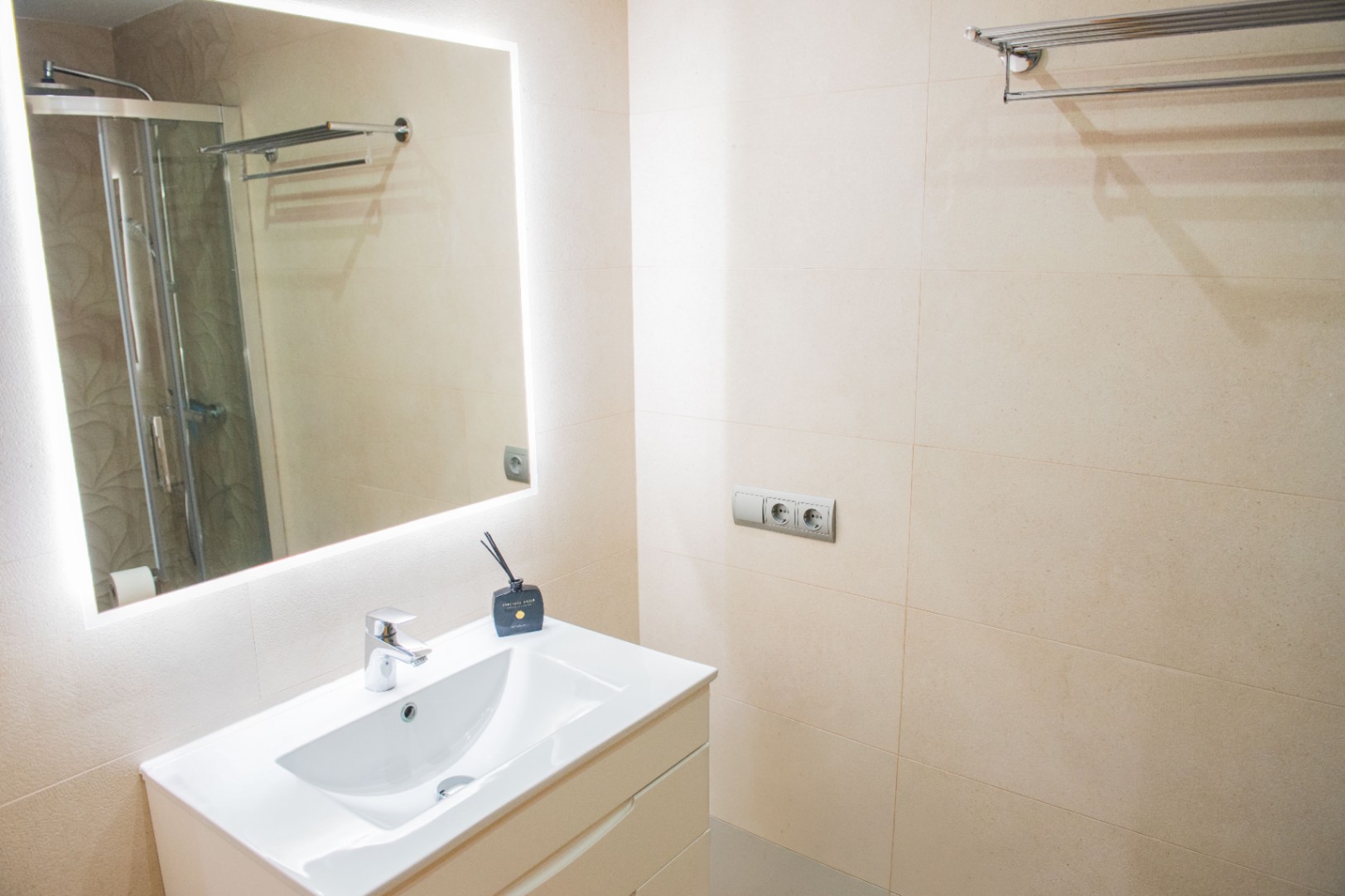 Modernes Badezimmer mit weißem Waschtisch, hintergrundbeleuchtetem LED-Spiegel und cremefarbenen Wandfliesen.