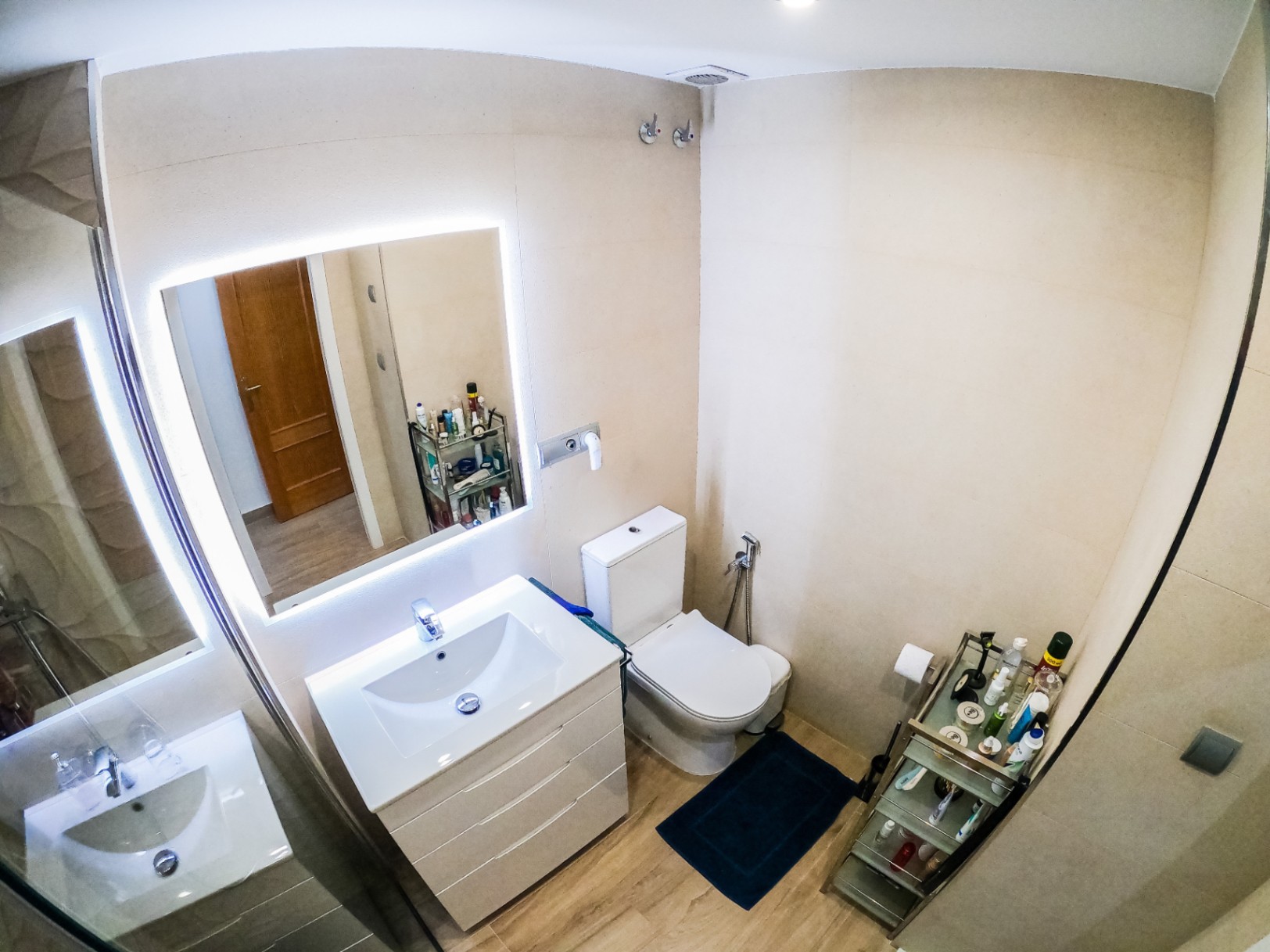 Modernes Badezimmer mit weißem Waschtisch, LED-hinterleuchtetem Spiegel und Boden in Holzoptik. Die Wände sind mit großformatigen beigen Fliesen gestaltet.