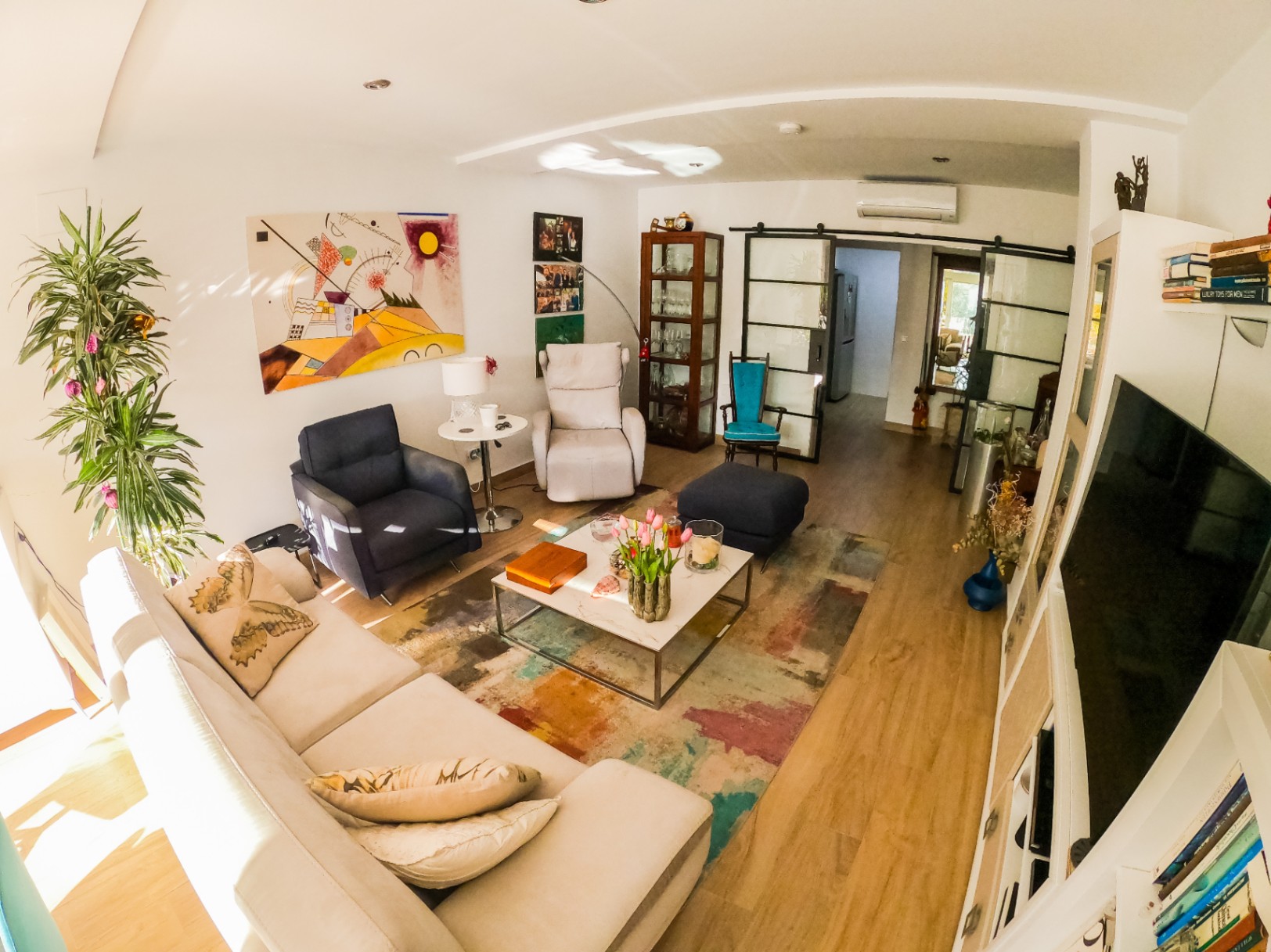 Modernes Wohnzimmer mit weißem Ecksofa, abstrakter Kunst und Marmorcouchtisch auf buntem Teppich. Industrielle Glasschiebetüren verbinden die Wohnbereiche.