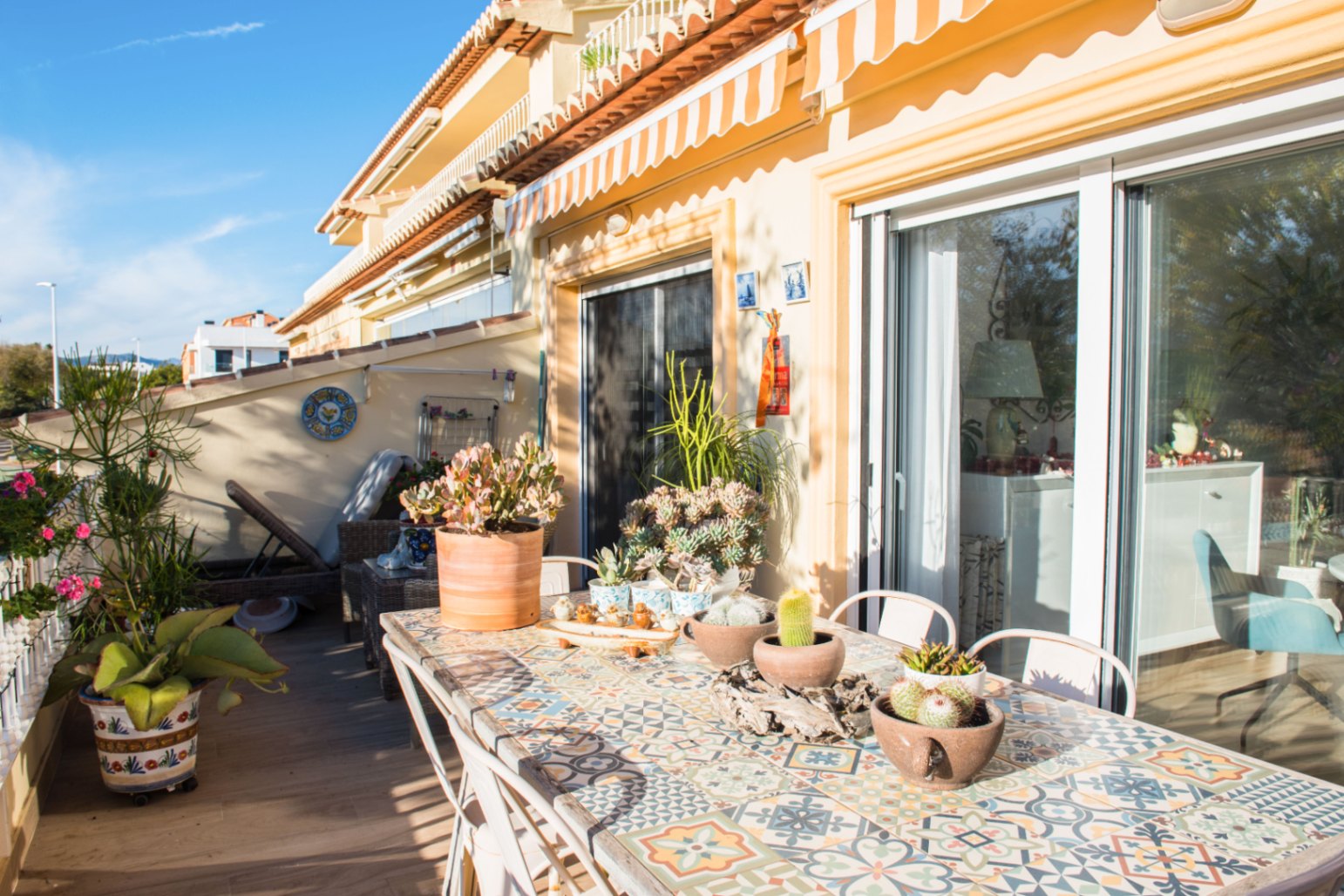 Sonnige mediterrane Terrasse mit einem Fliesentisch im Mosaik-Stil, Sukkulenten und großen Glasschiebetüren unter einer gestreiften Markise.