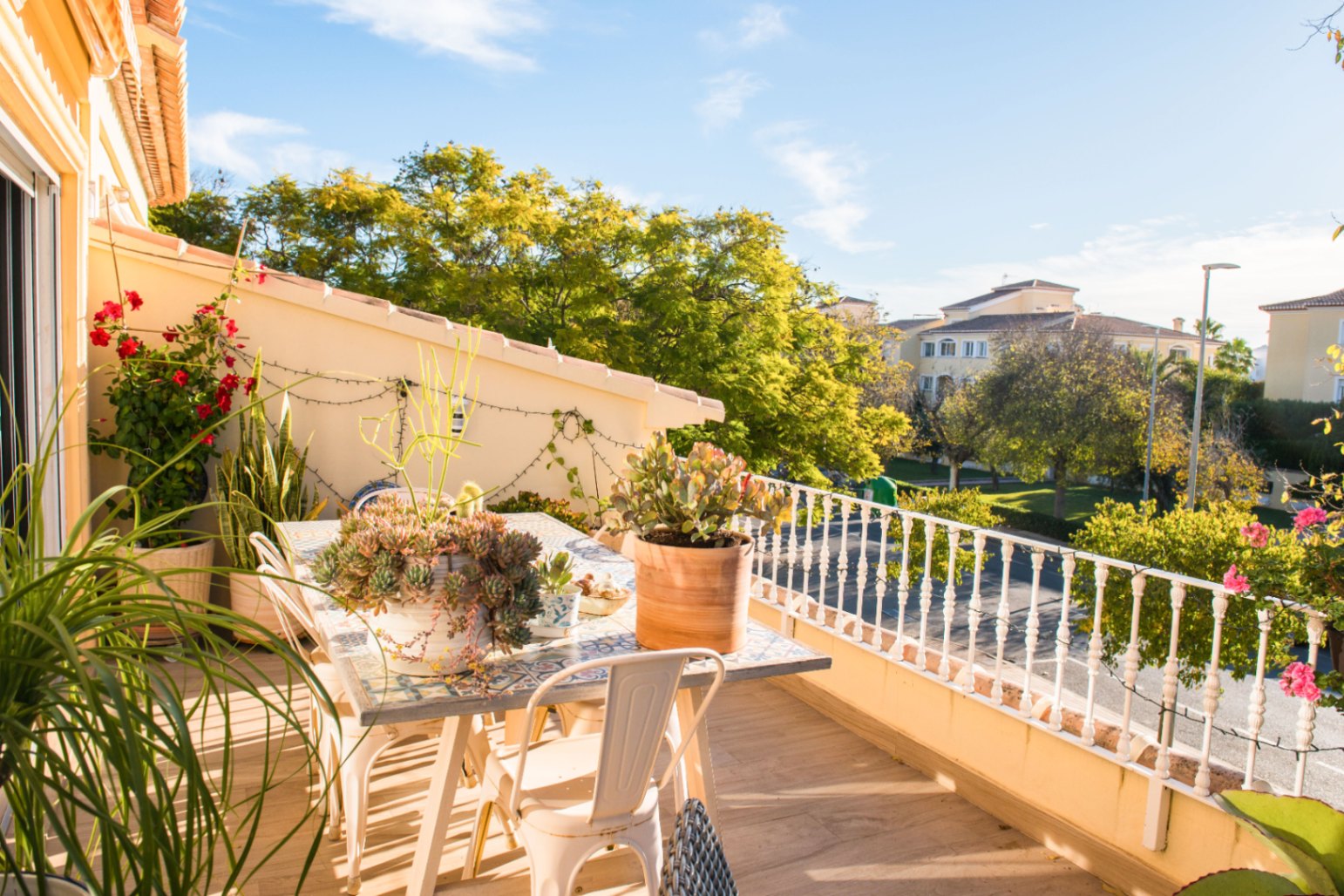 Terrasse mit Mosaik-Esstisch, weißen Metallstühlen und Sukkulenten in Töpfen mit Blick auf eine Wohngegend und ausgewachsene Bäume.