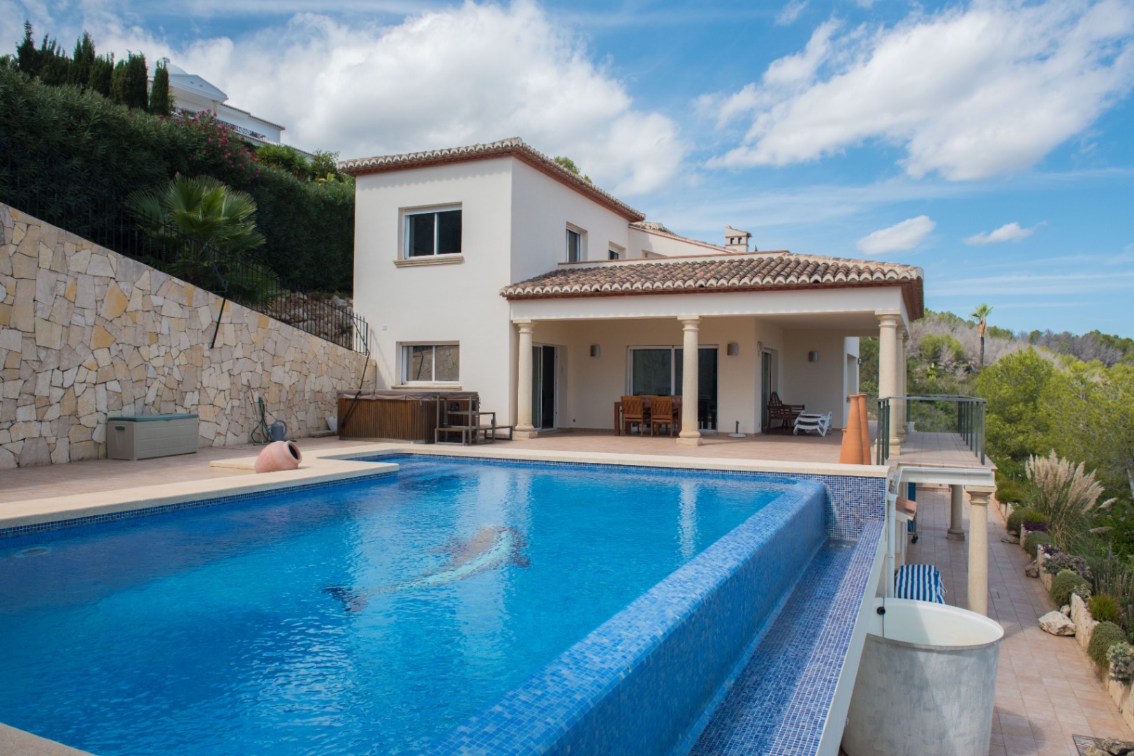 Außenansicht einer weißen Villa mit Infinity-Pool aus blauem Mosaik, Delfin-Motiv und einer überdachten Terrasse mit Säulen und Natursteinwand.