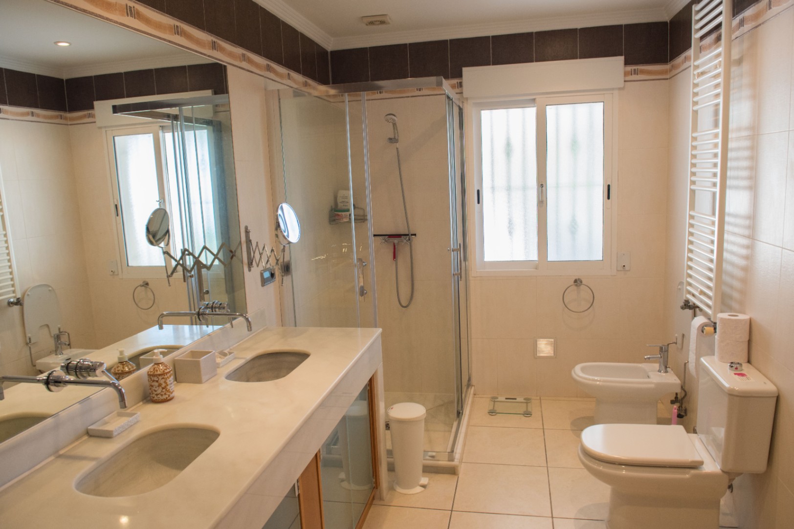 Badezimmer mit Doppelwaschtisch aus Marmor, einer gläsernen Eckdusche sowie WC und Bidet, eingefasst in helle Wandfliesen mit dekorativer Bordüre.