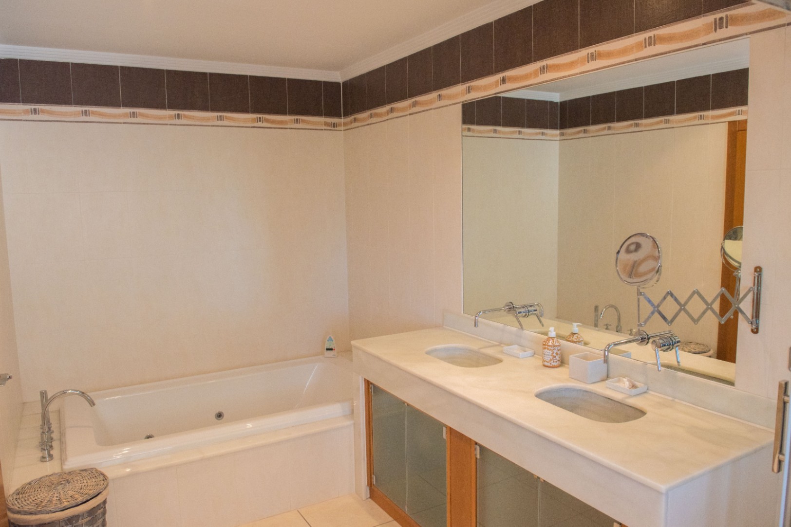 Badezimmer mit Doppelwaschtisch aus Stein, Wandarmaturen und eingebauter Whirlpool-Badewanne, ergänzt durch helle Wandfliesen mit dekorativer Bordüre.