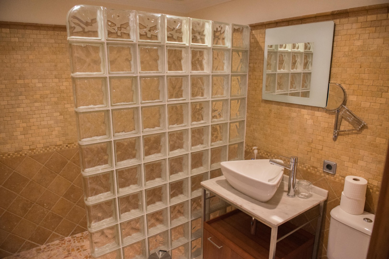 Badezimmer mit einer Duschwand aus Glasbausteinen, beigen Mosaikfliesen und einem Aufsatzwaschbecken auf einer Marmorplatte mit Holzunterschrank.