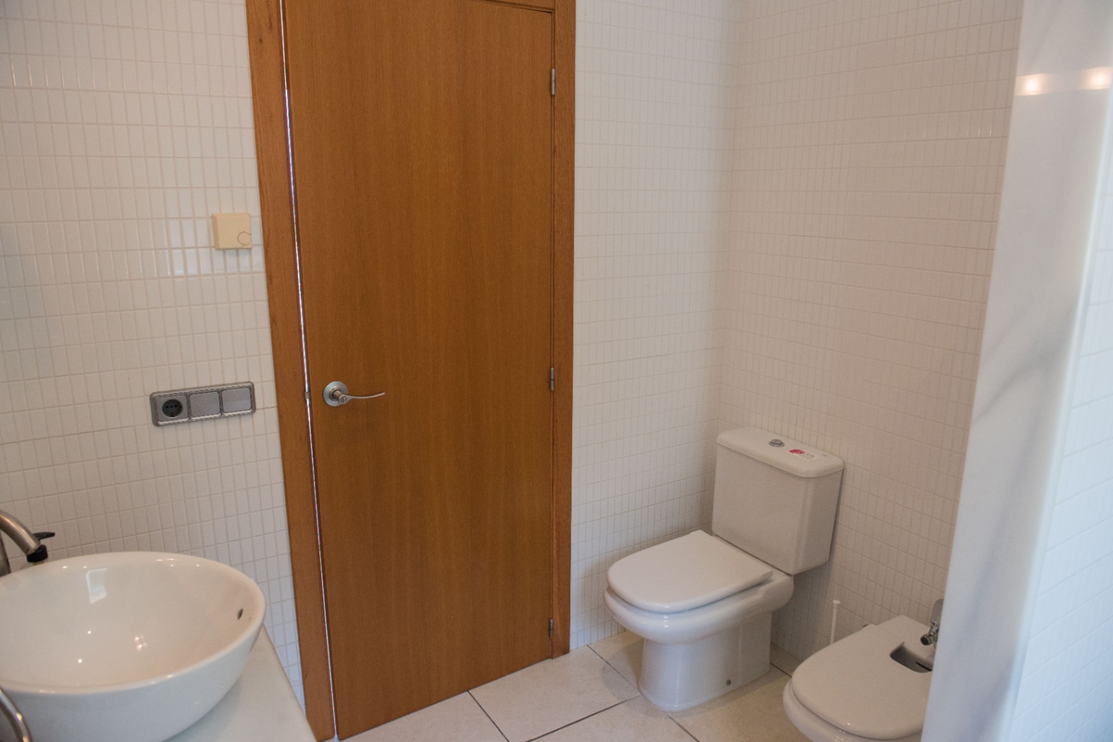 Badezimmer mit weißen quadratischen Wandfliesen, Holztür, Keramik-WC, bidet und einem weißen Aufsatzwaschbecken mit Chromarmaturen.