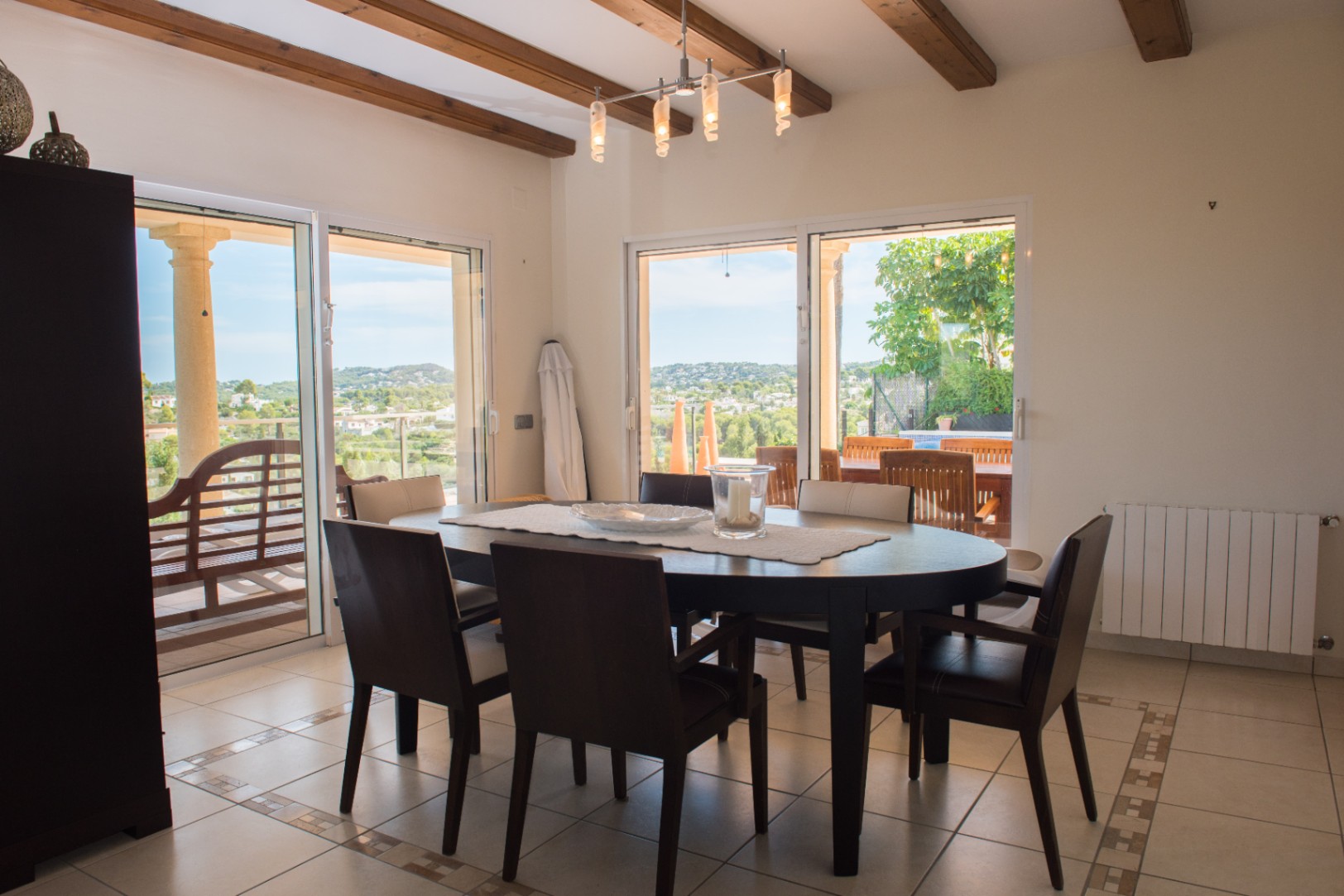 Essbereich mit ovalem dunklem Holztisch, sichtbaren Deckenbalken und großen Glasschiebetüren, die einen weiten Blick auf die Hügel und Zugang zur Terrasse bieten.