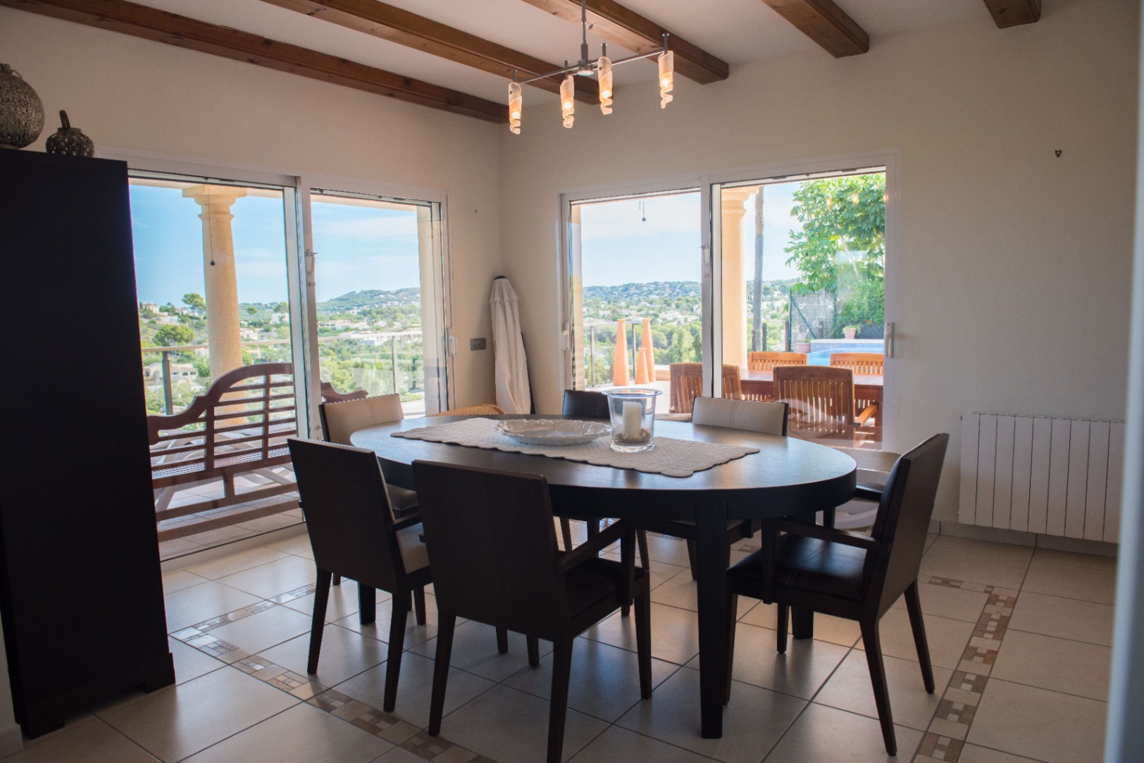 Essbereich mit ovalem dunklem Holztisch und sichtbaren Deckenbalken. Große Glasschiebetüren bieten einen Panoramablick über das Tal und direkten Zugang zur Terrasse.