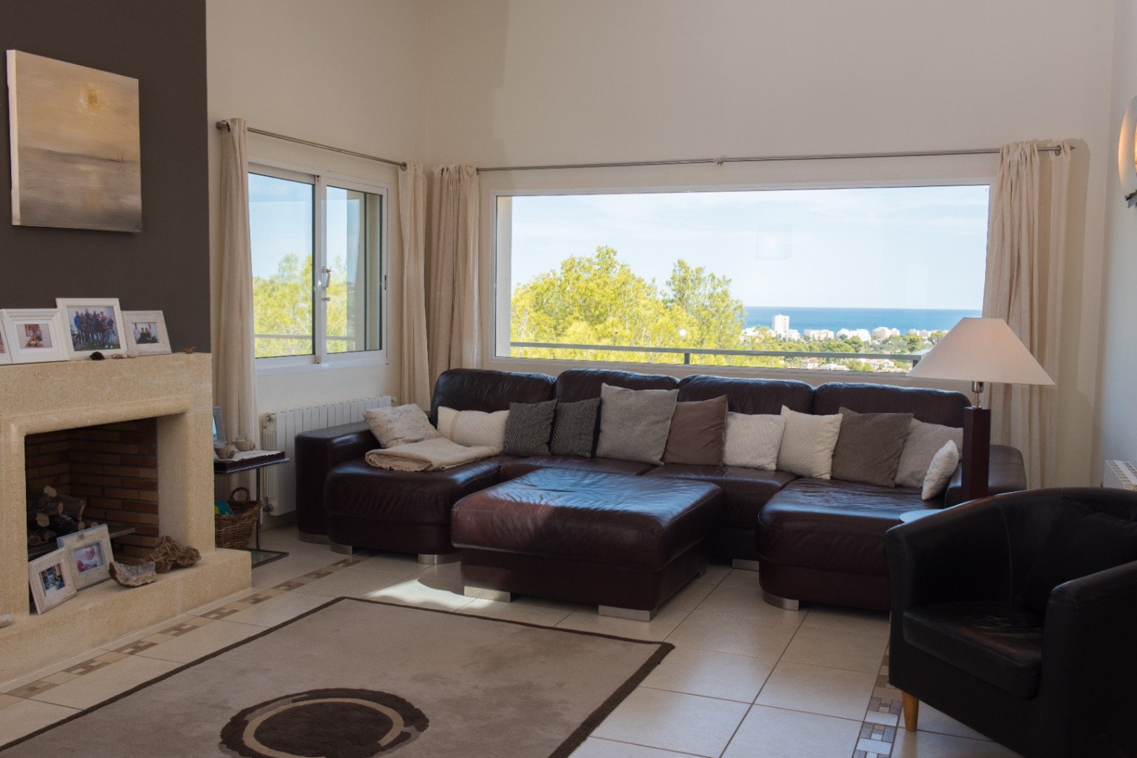 Großzügiges Wohnzimmer mit brauner Ledereckcouch, Natursteinkamin und einem großen Panoramafenster mit Blick auf das Meer und die Küstenlandschaft.