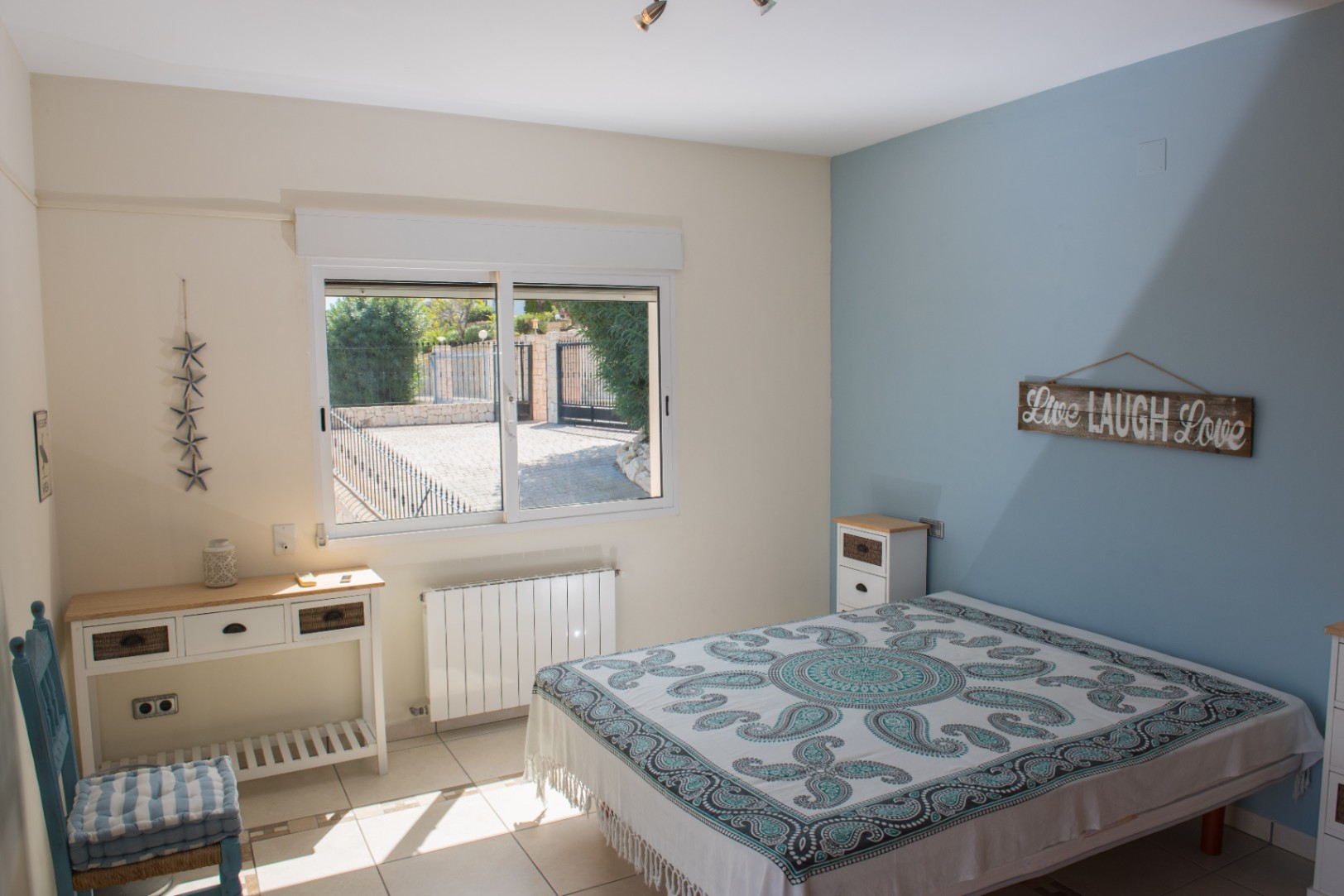 Helles Schlafzimmer mit blauer Akzentwand, Fliesenboden und Fenster mit Blick auf die gepflasterte Einfahrt. Ausgestattet mit weißem Konsolentisch und Heizkörper.