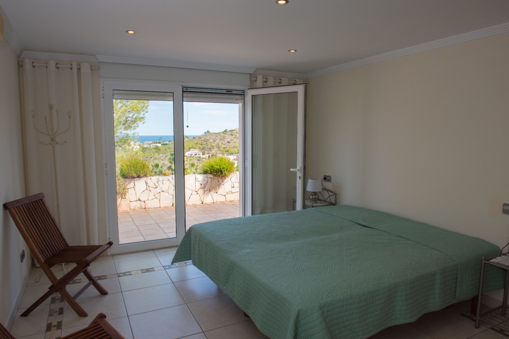 Helles Schlafzimmer mit direktem Zugang zur Terrasse und Blick auf das Meer und die Hügel, ausgestattet mit Fliesenboden und dezenten Deckenstrahlern.