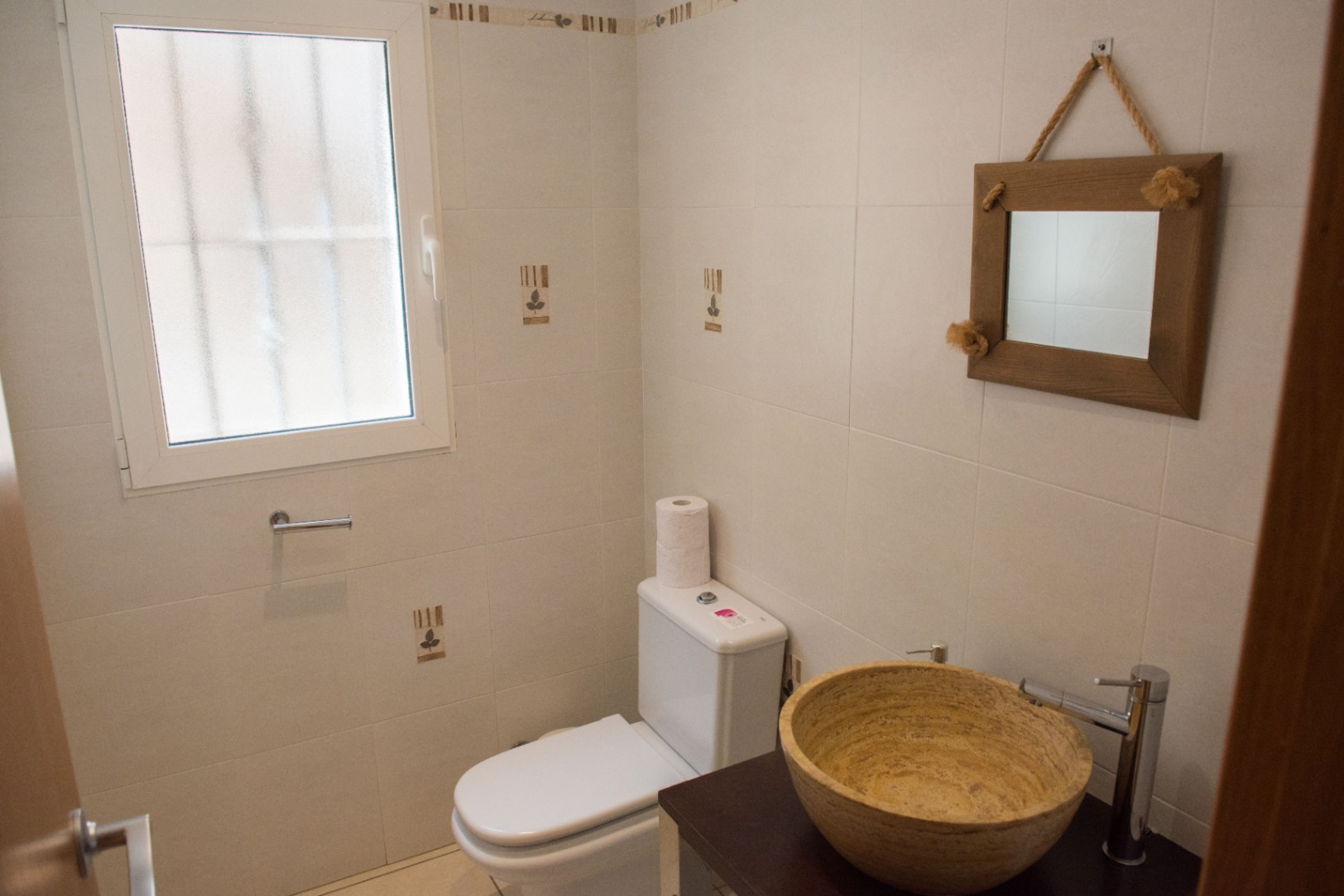 Kompaktes Gäste-WC mit Naturstein-Aufsatzwaschbecken auf dunklem Holzwaschtisch, rustikalem Seilspiegel und hell gefliesten Wänden mit mattiertem Fenster.