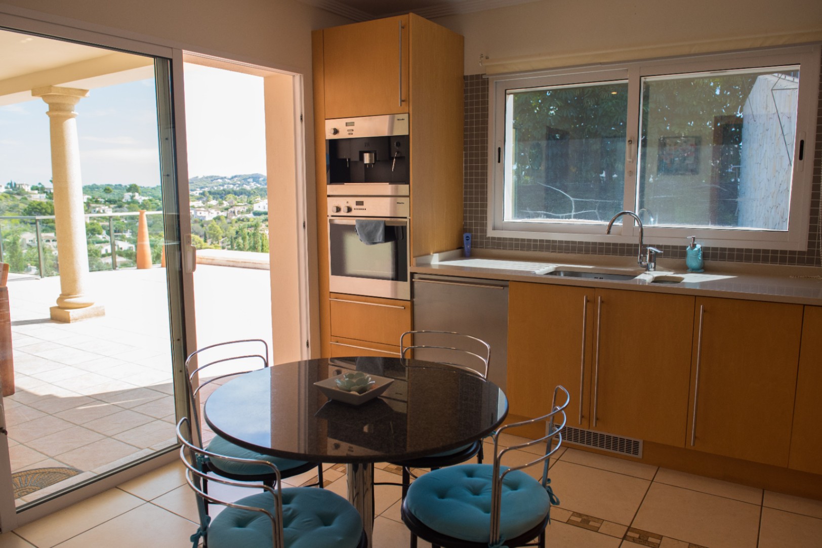 Küche mit hellen Holzfronten, integrierten Miele-Geräten und Granit-Esstisch. Eine Glasschiebetür führt zur Terrasse mit Panoramablick über das Tal.