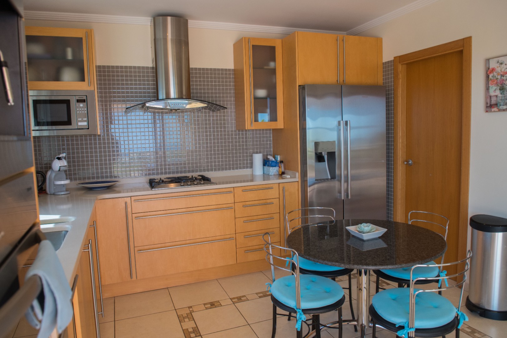 Moderne Küche mit hellen Holzschränken, grauem Mosaik-Fliesenspiegel und Edelstahlgeräten. Ein runder Granittisch mit blau gepolsterten Stühlen ergänzt den Raum.