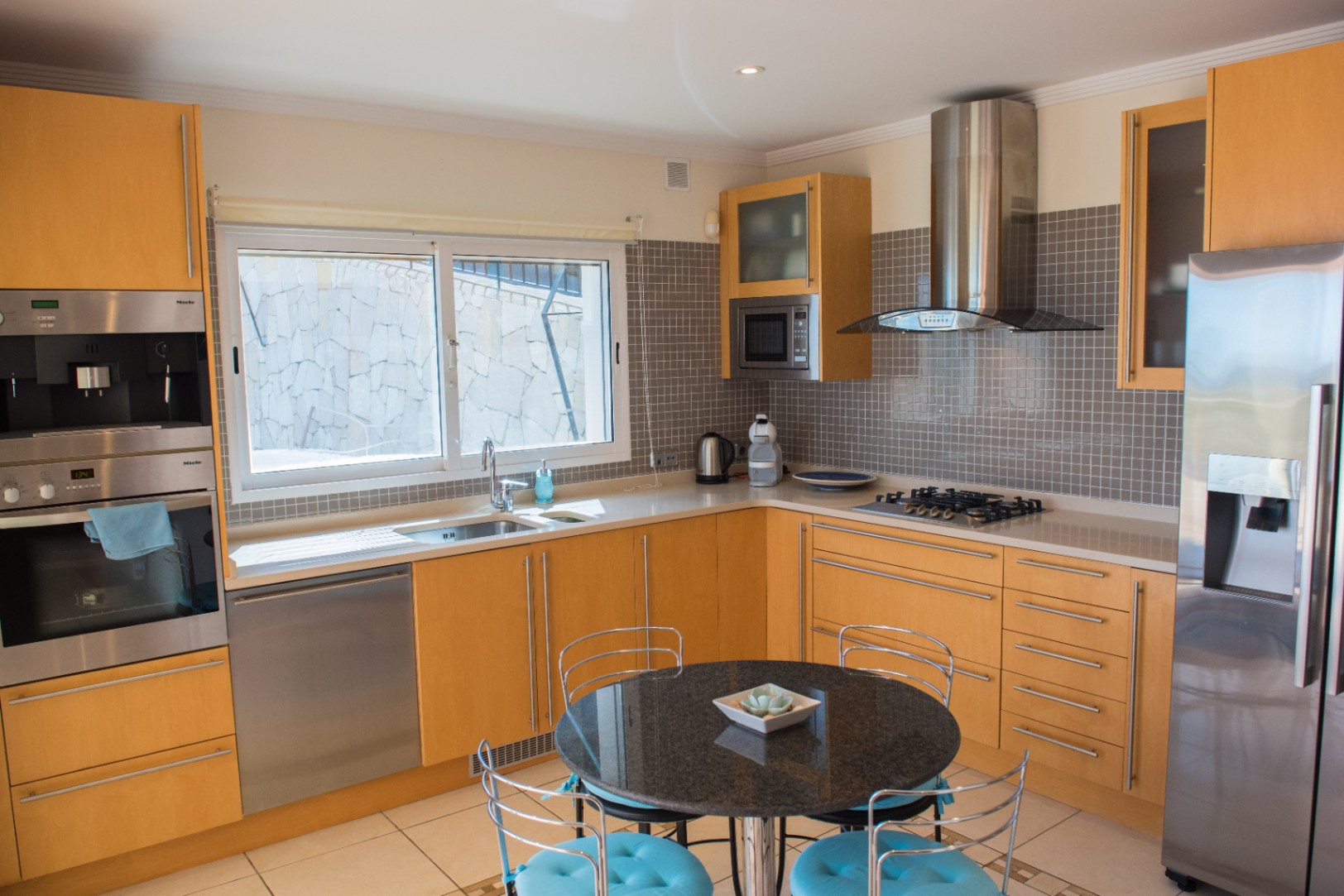 Moderne Küche mit hellen Holzschränken, Miele-Edelstahlgeräten und grauem Mosaik-Fliesenspiegel. Mit rundem Granit-Frühstückstisch und integriertem Gaskochfeld.