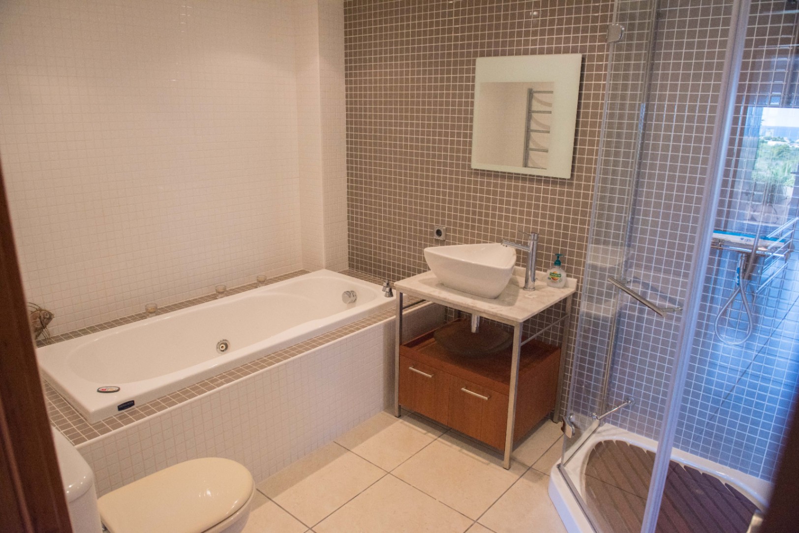 Modernes Badezimmer mit Einbauwanne, Glasdusche mit Holzeinlage und Aufsatzwaschbecken auf Marmorplatte, akzentuiert durch taupefarbene Mosaikfliesen.