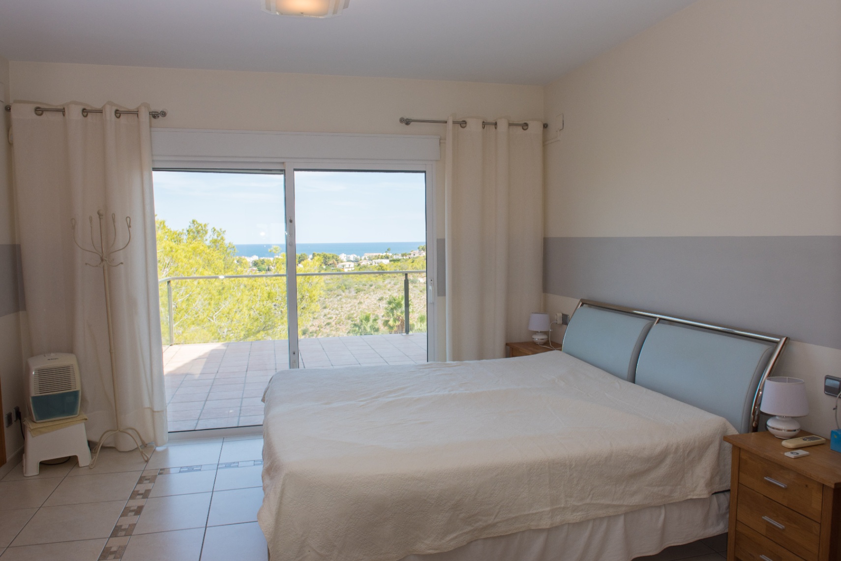 Schlafzimmer mit Fliesenboden und Glasschiebetüren zur Terrasse mit Meerblick. Ausgestattet mit einem modernen Metallkopfteil und neutralen Wandfarben.