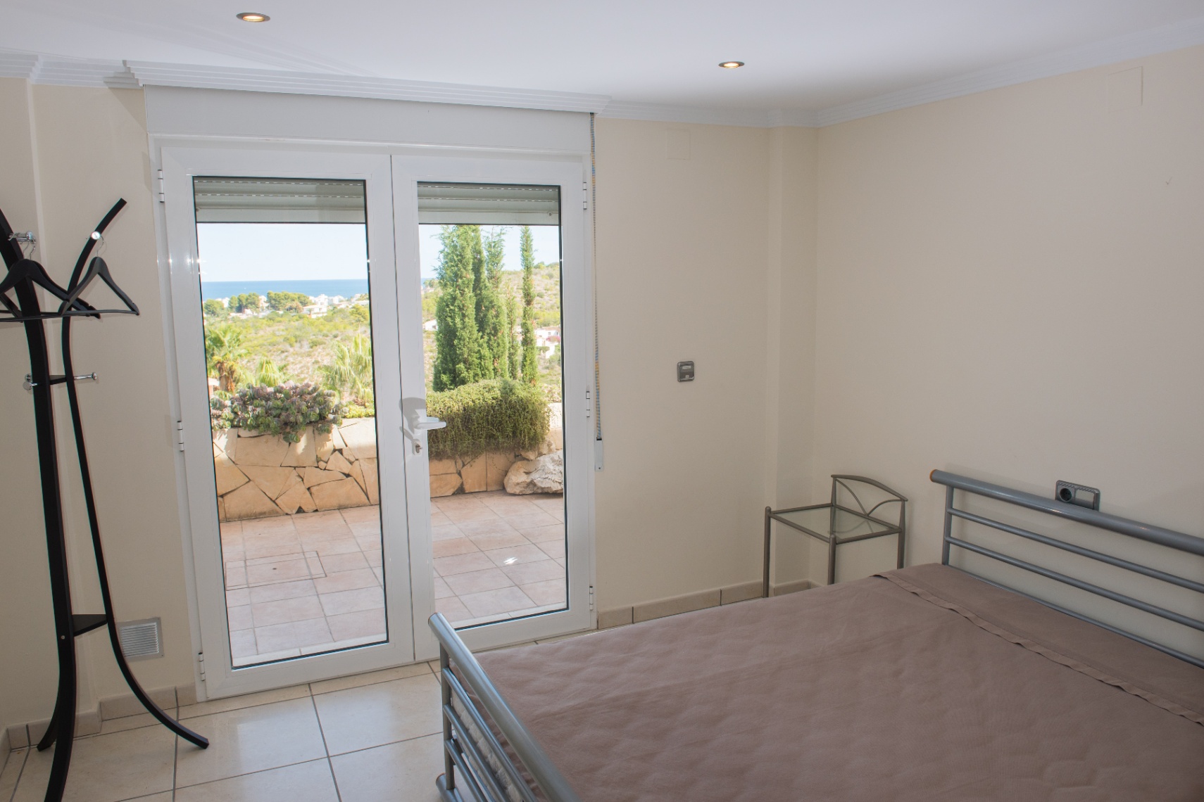 Schlafzimmer mit grauem Metallbettrahmen und Glastisch, mit direktem Zugang zu einer gefliesten Terrasse mit Blick auf die Küste.