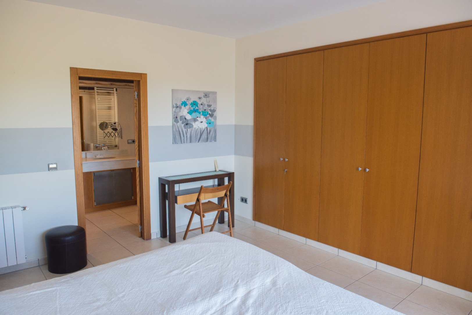 Schlafzimmer mit großem Einbauschrank aus Holz, kleinem Arbeitsbereich und angrenzendem Badezimmer mit hellen Fliesen.