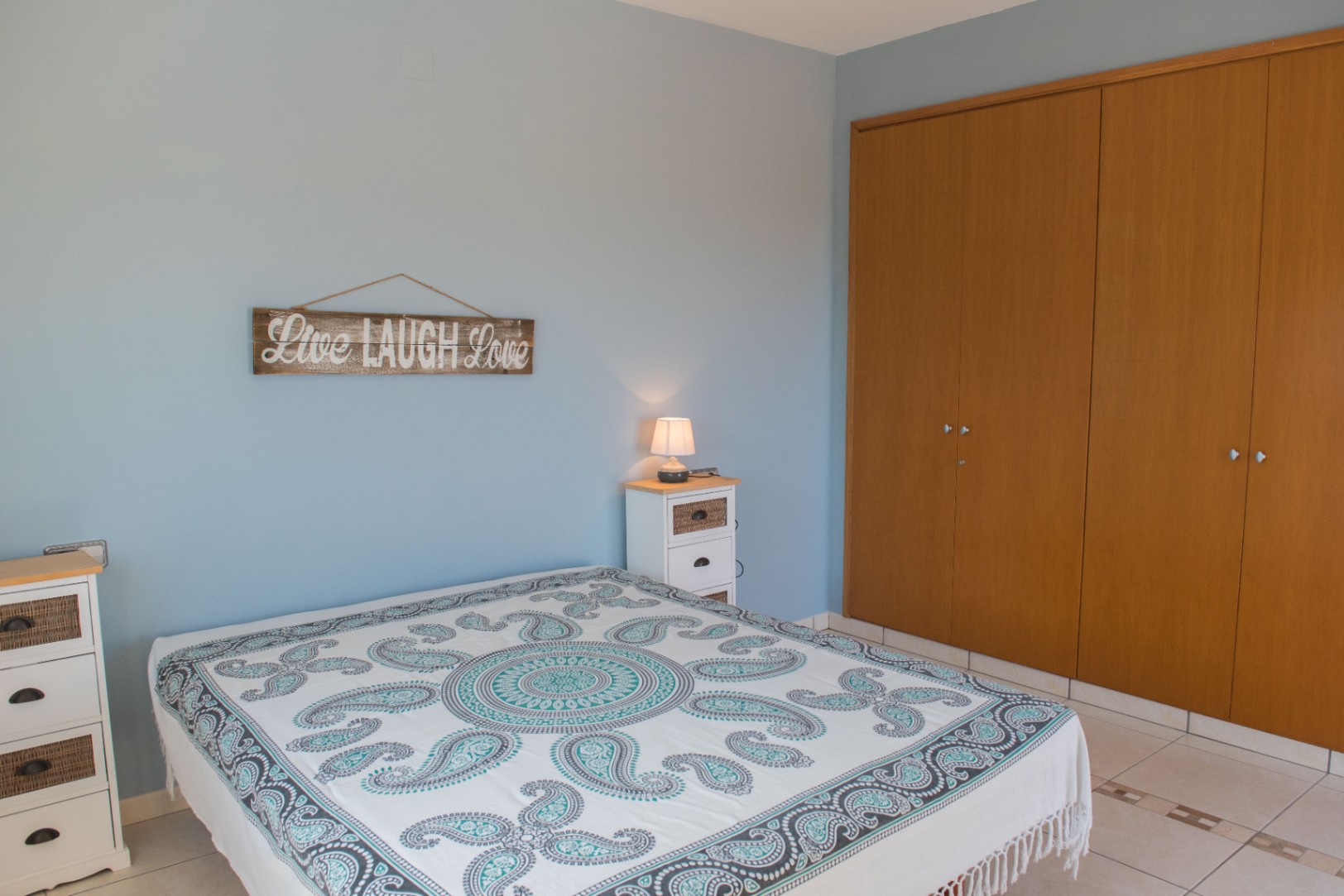 Schlafzimmer mit hellblauen Wänden, einem großen Holz-Einbauschrank und weißen Möbeln mit Weidendetails. Der Raum verfügt über einen Fliesenboden und eine gemusterte Decke.