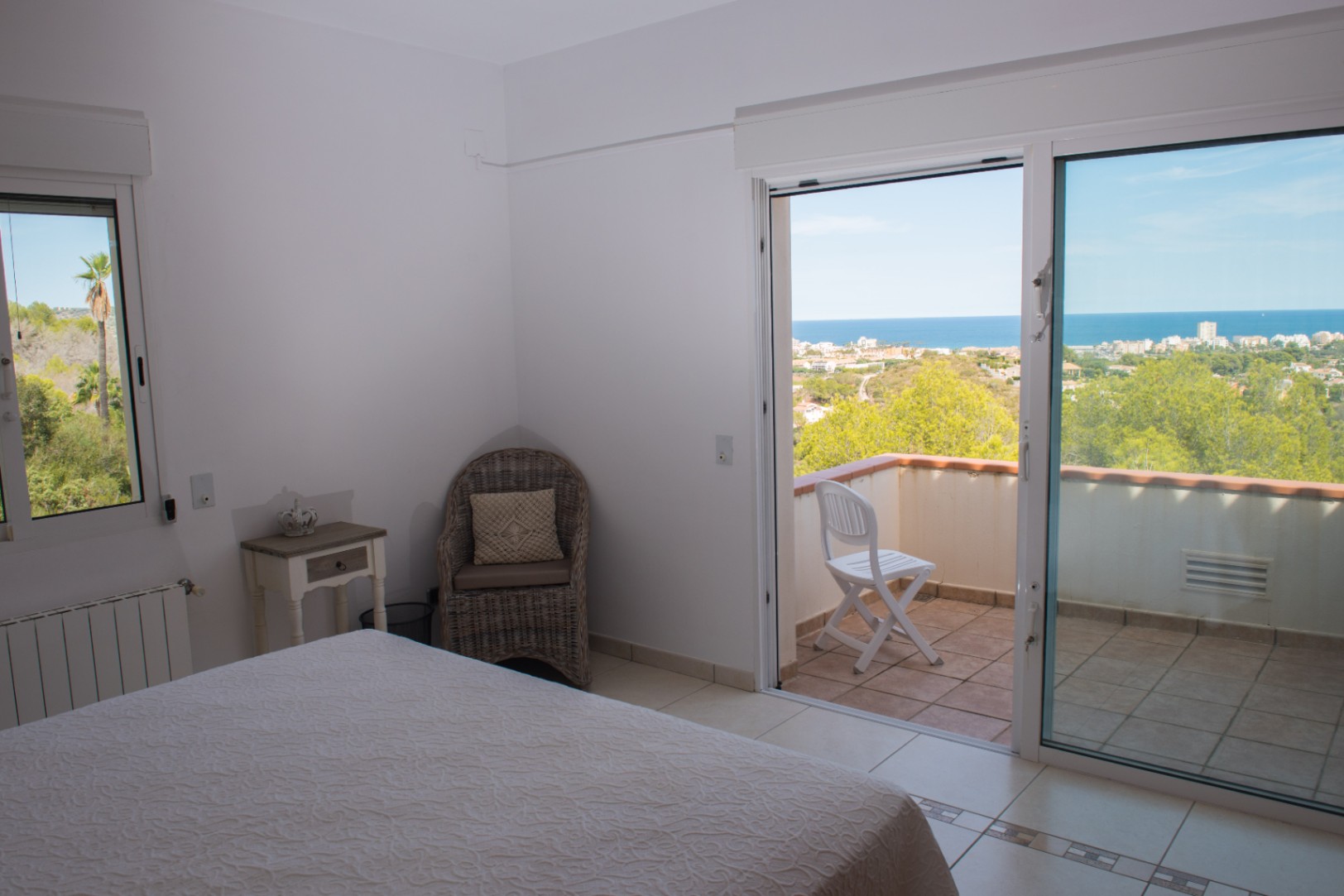 Schlafzimmer mit Zugang zum Balkon und Panoramablick auf das Meer. Ausgestattet mit hellen Fliesen, einem Korbsessel und einer großen Glasschiebetür.