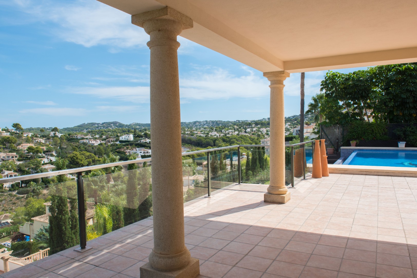 Überdachte Terrasse mit Steinsäulen und Glasgeländer, die einen Panoramablick über das Tal und den Poolbereich bietet.