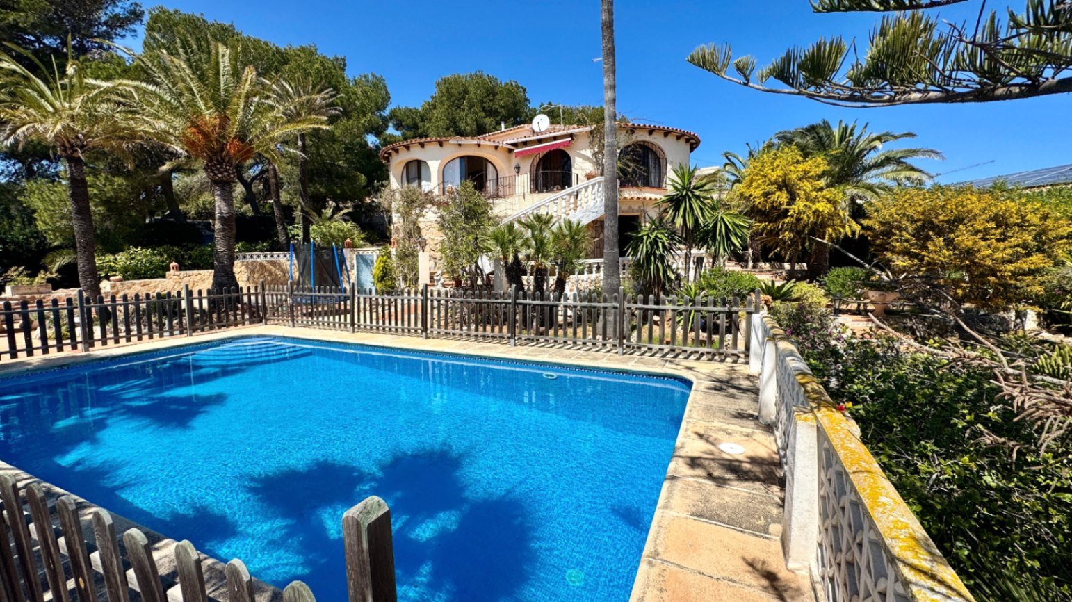 Außenansicht mit umzäuntem rechteckigem Pool und einer mediterranen Villa mit Rundbogenbalkonen, eingebettet in einen Garten mit Palmen.