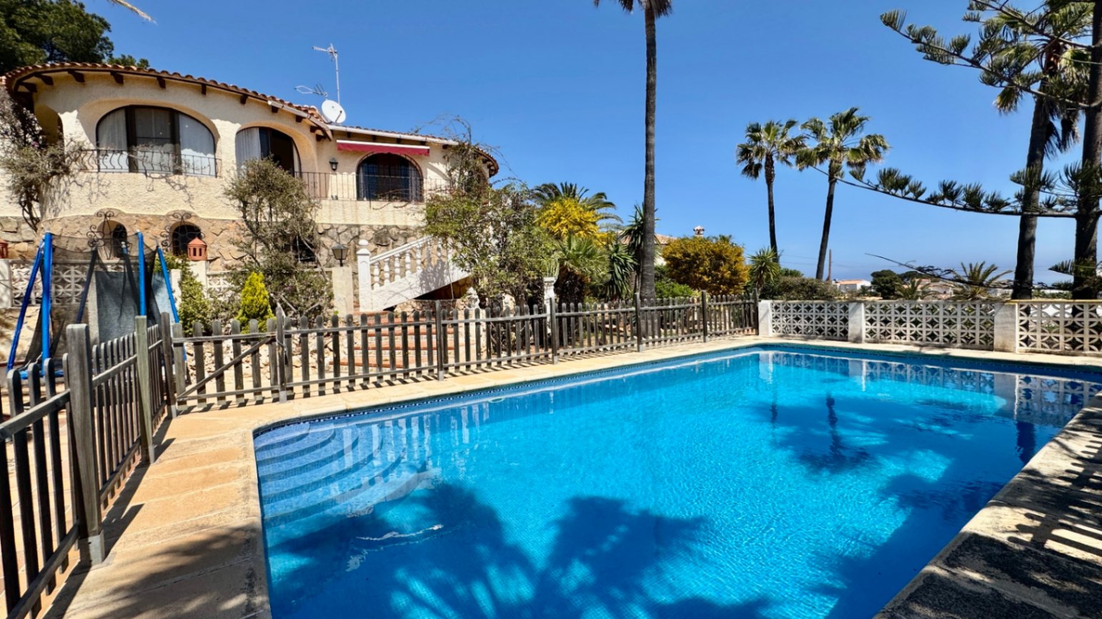 Außenpool mit blauen Fliesen und integrierter Treppe, umgeben von einem Holzzaun und einer mediterranen Villa mit Steinfassade und Rundbogenbalkonen.