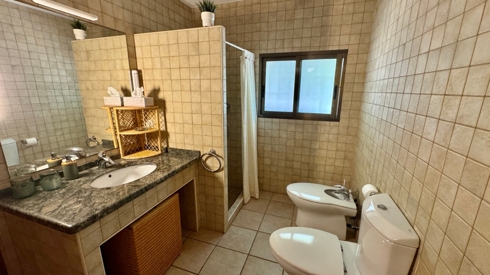 Badezimmer mit Granit-Waschtisch, integriertem Waschbecken und Korb-Accessoires. Beige Wandfliesen, ebenerdige Dusche mit Vorhang sowie WC und Bidet.