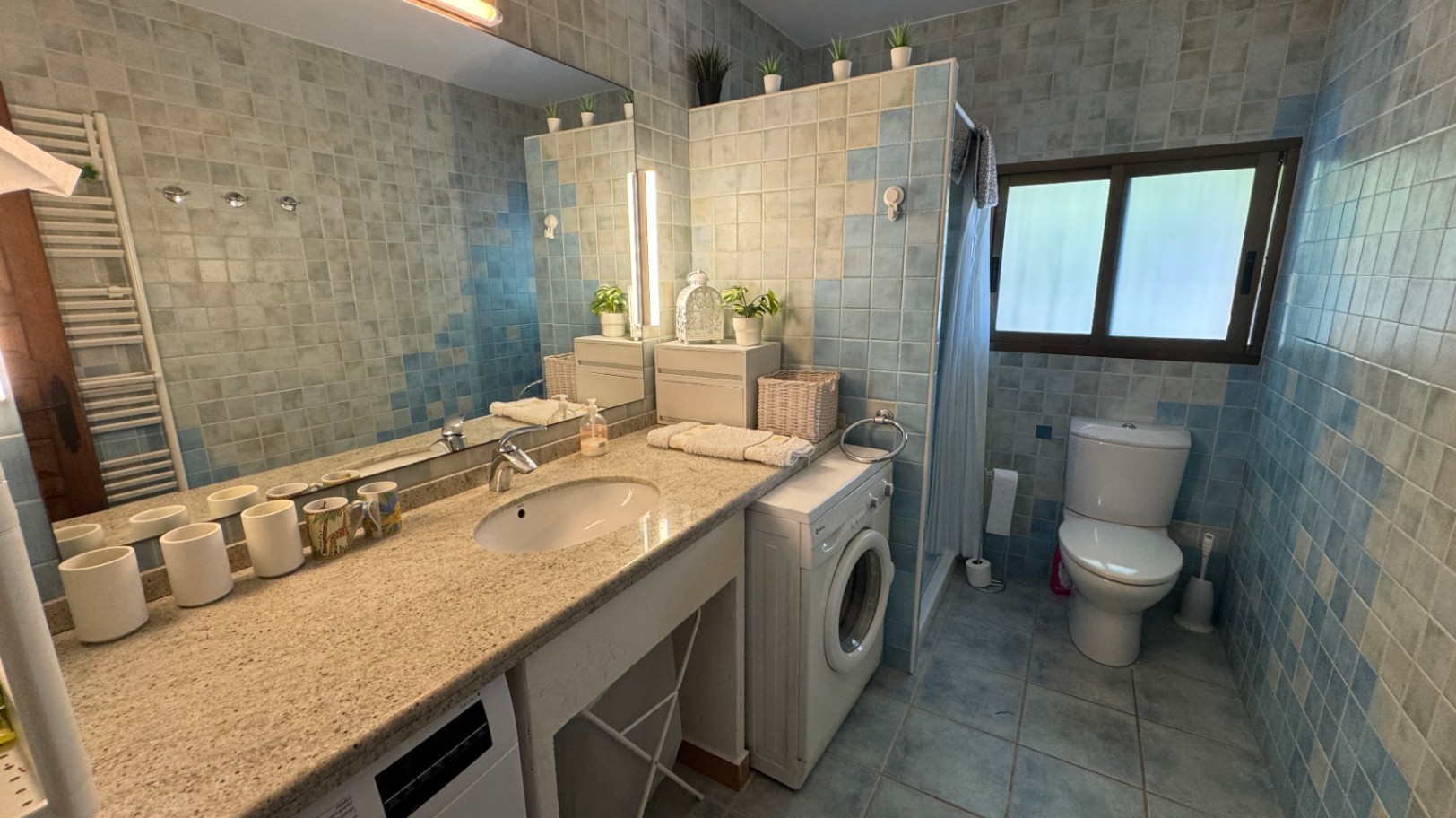 Badezimmer mit Granitwaschtisch, blauen Mosaikfliesen und integrierter Waschmaschine. Mit großem Spiegel, Beleuchtung und Außenfenster.