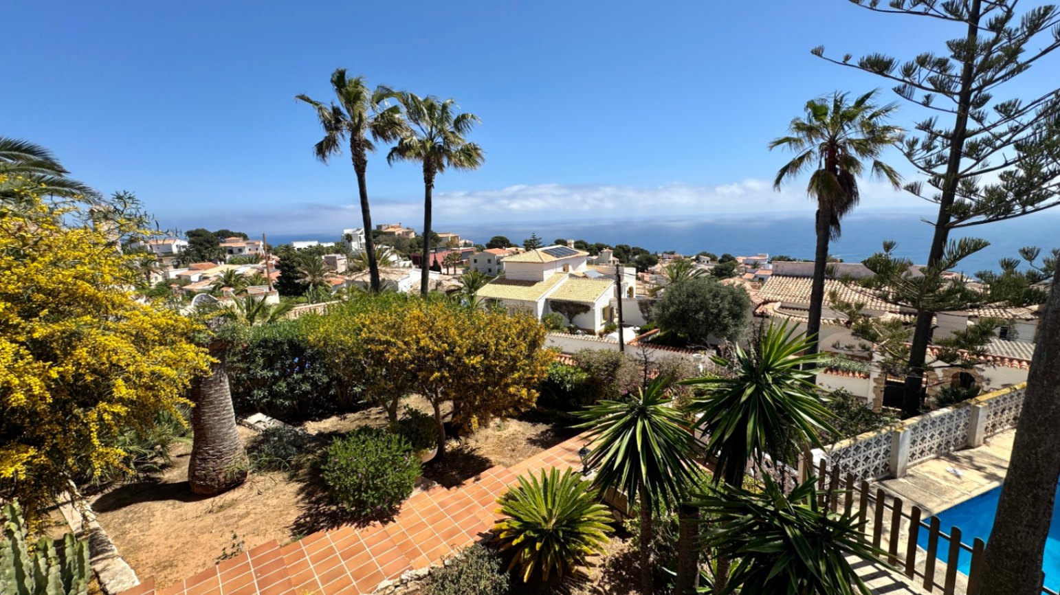 Erhöhter Blick auf einen mediterranen Garten mit Terrakottawegen und Palmen, mit Aussicht auf ein Küstenwohngebiet und das offene Meer.
