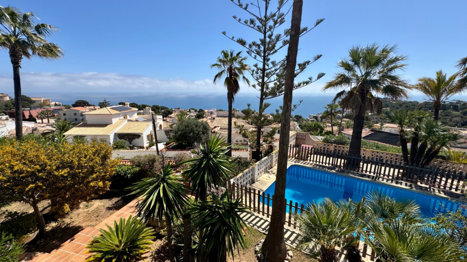 Erhöhter Blick auf einen privaten Pool mit Holzzaun, umgeben von Palmen und mediterranen Gärten mit weitem Blick auf das Meer.