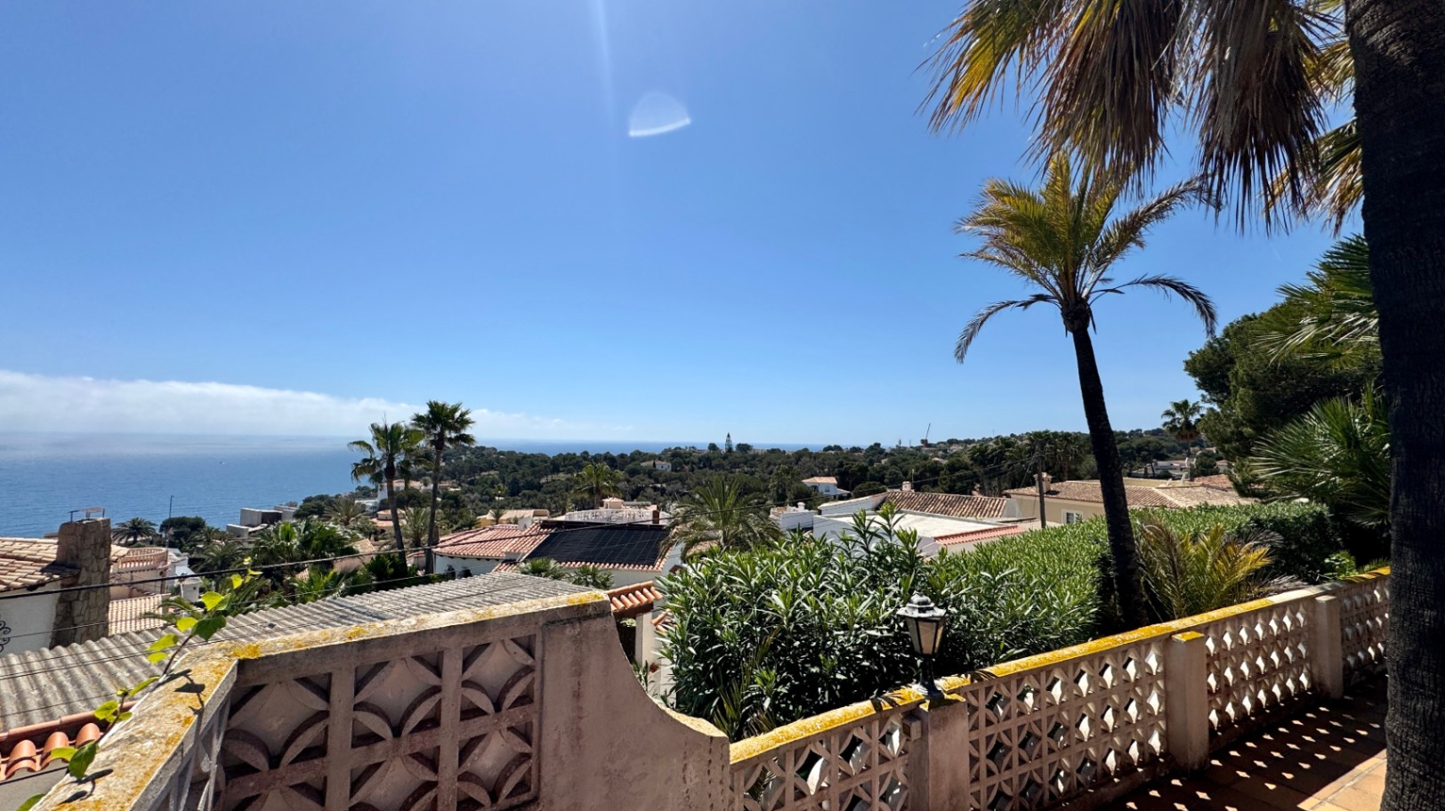 Erhöhter Terrassenblick über eine mediterrane Siedlung mit Terrakottadächern, Palmen und weitem Meerblick am Horizont unter klarem blauem Himmel.