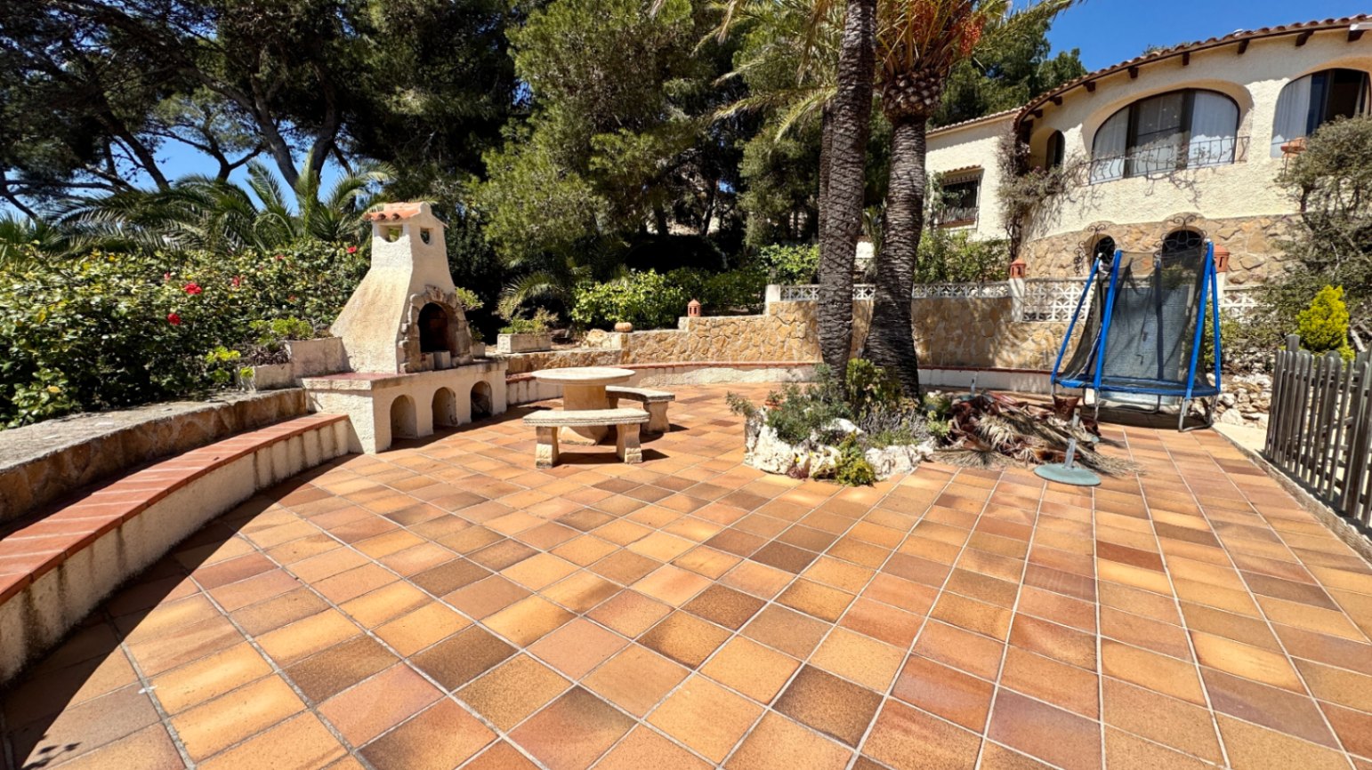 Großzügige Terrasse mit Terrakotta-Fliesen, gemauertem Steingrill, Steinsitzgruppe und Palmen vor einer mediterranen Villa.