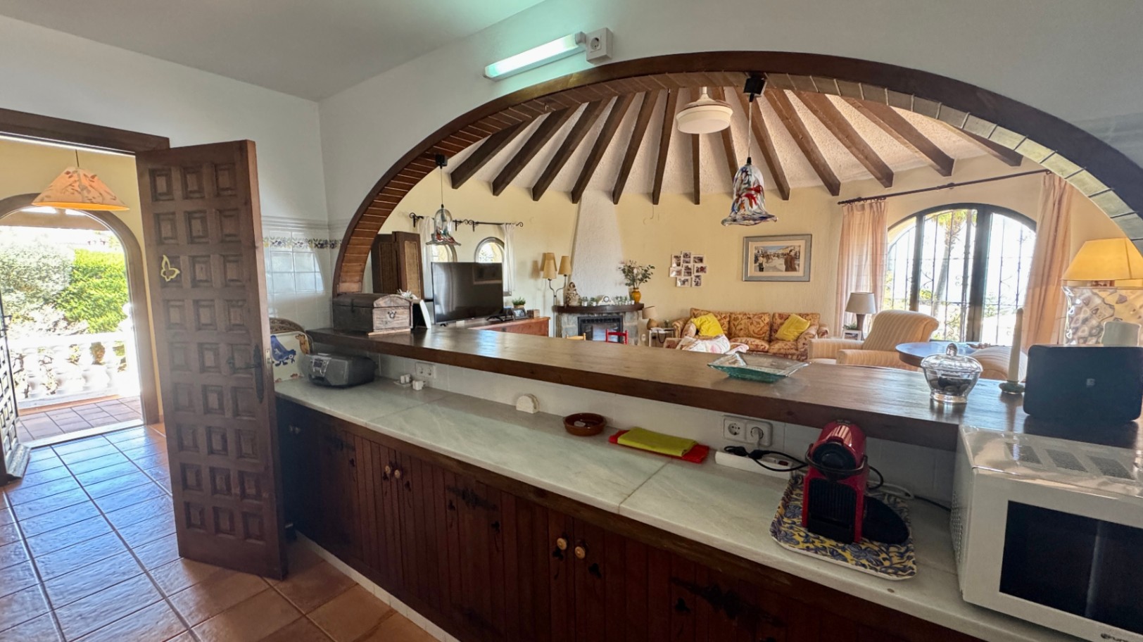 Küchentheke mit Marmorplatte und Holzschränken, die durch einen großen Holzbogen in ein rundes Wohnzimmer mit radialen Deckenbalken blickt.