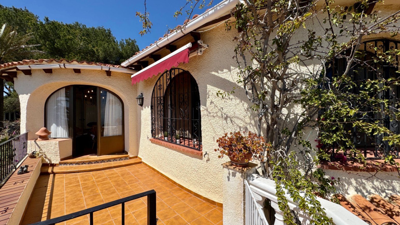 Mediterrane Villa mit Terrakotta-Terrasse, Rundbogenfenstern mit schmiedeeisernen Gittern und einer roten Markise vor hellen Putzwänden.