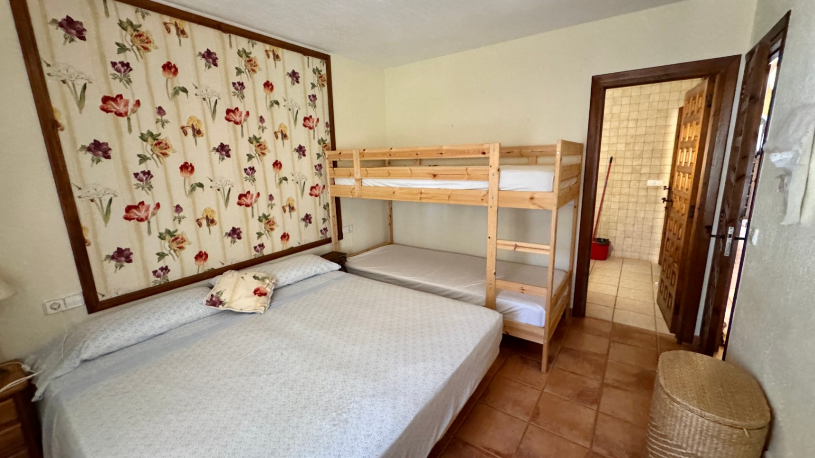 Schlafzimmer mit Doppelbett und Holz-Etagenbett, ergänzt durch ein florales Wandpaneel und Terrakotta-Fliesen. Ein angrenzendes Bad ist durch die Tür sichtbar.